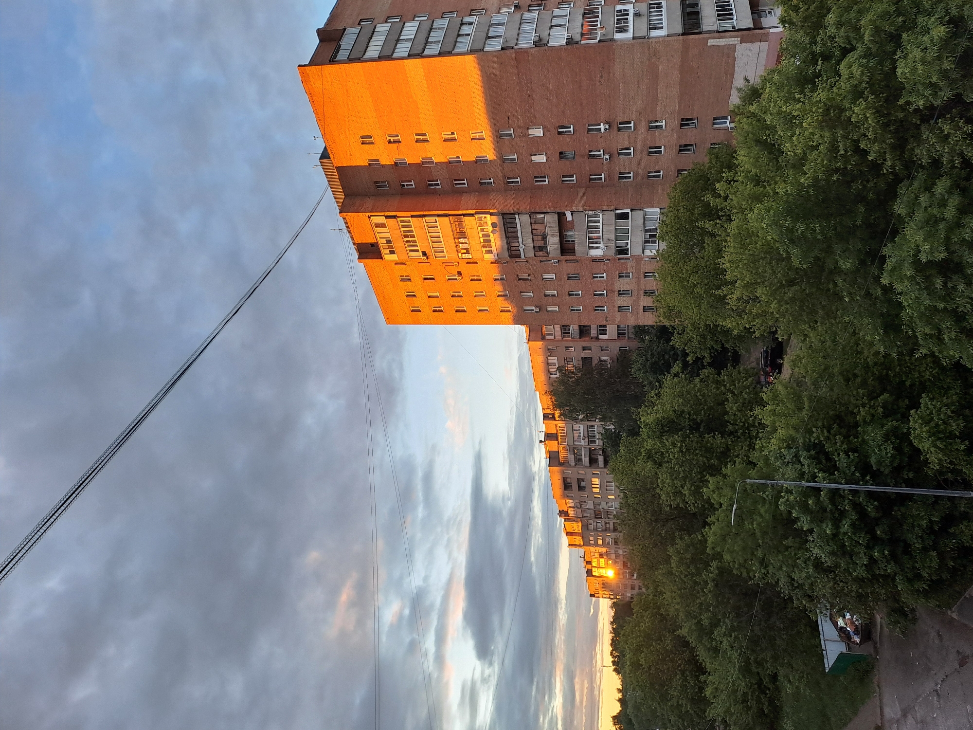 A low-angle shot reveals a cityscape at dusk in Elektrostal, Russia. On the right, a brick apartment building dominates the view, its facade illuminated by the setting sun. A power line stretches across the upper left portion of the image. Lush green trees line the street. A street light is visible among the trees. The sky is filled with clouds, tinged with the colors of the sunset.