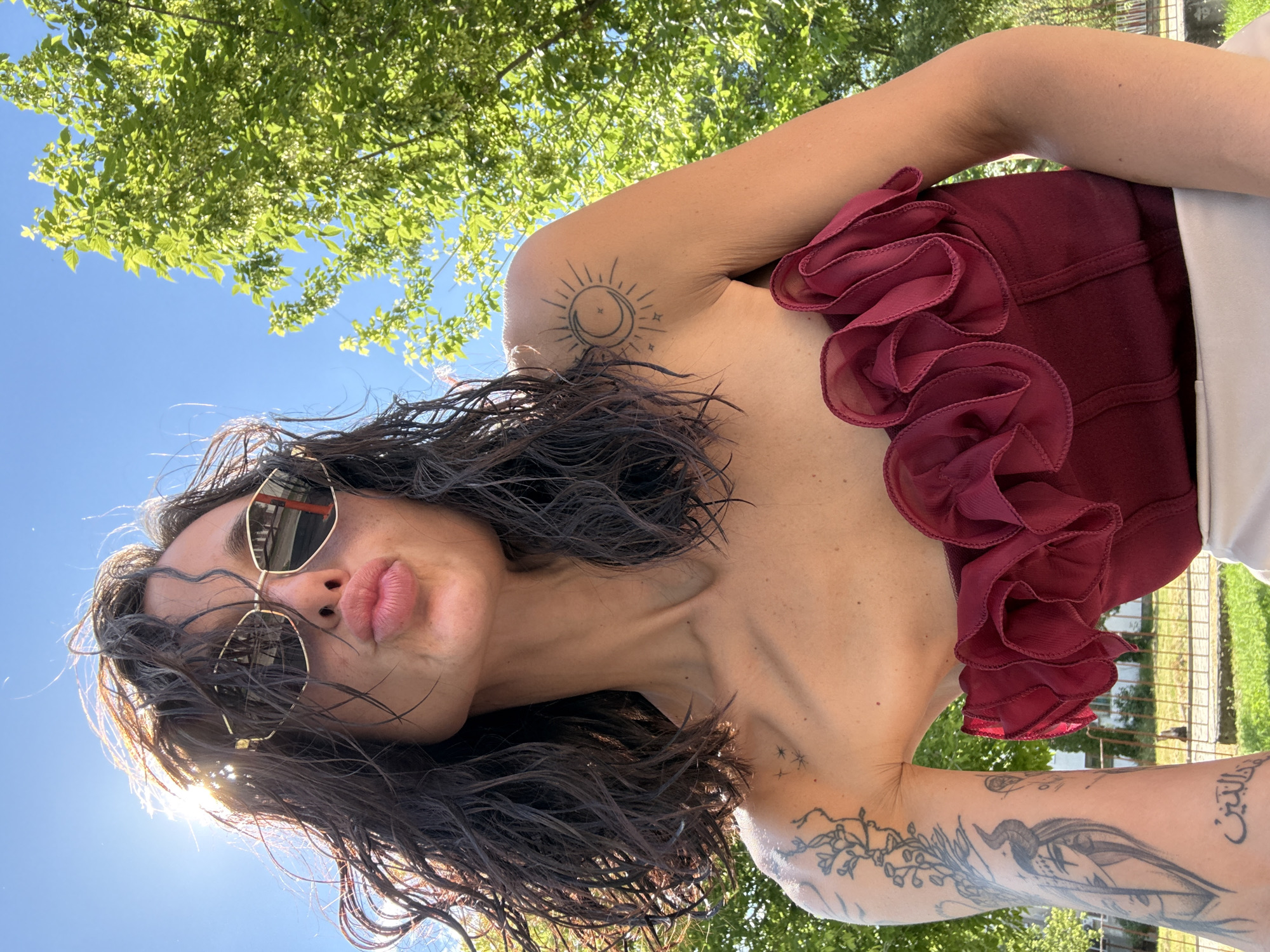 A woman is taking a selfie outdoors on a sunny day. She has dark, wet hair and is wearing sunglasses, a strapless maroon top with ruffles, and a light-colored skirt. She is making a kissing face. Tattoos are visible on her left arm and shoulder. The background shows green trees and a bright blue sky. The location is the city of Pleven, Bulgaria.