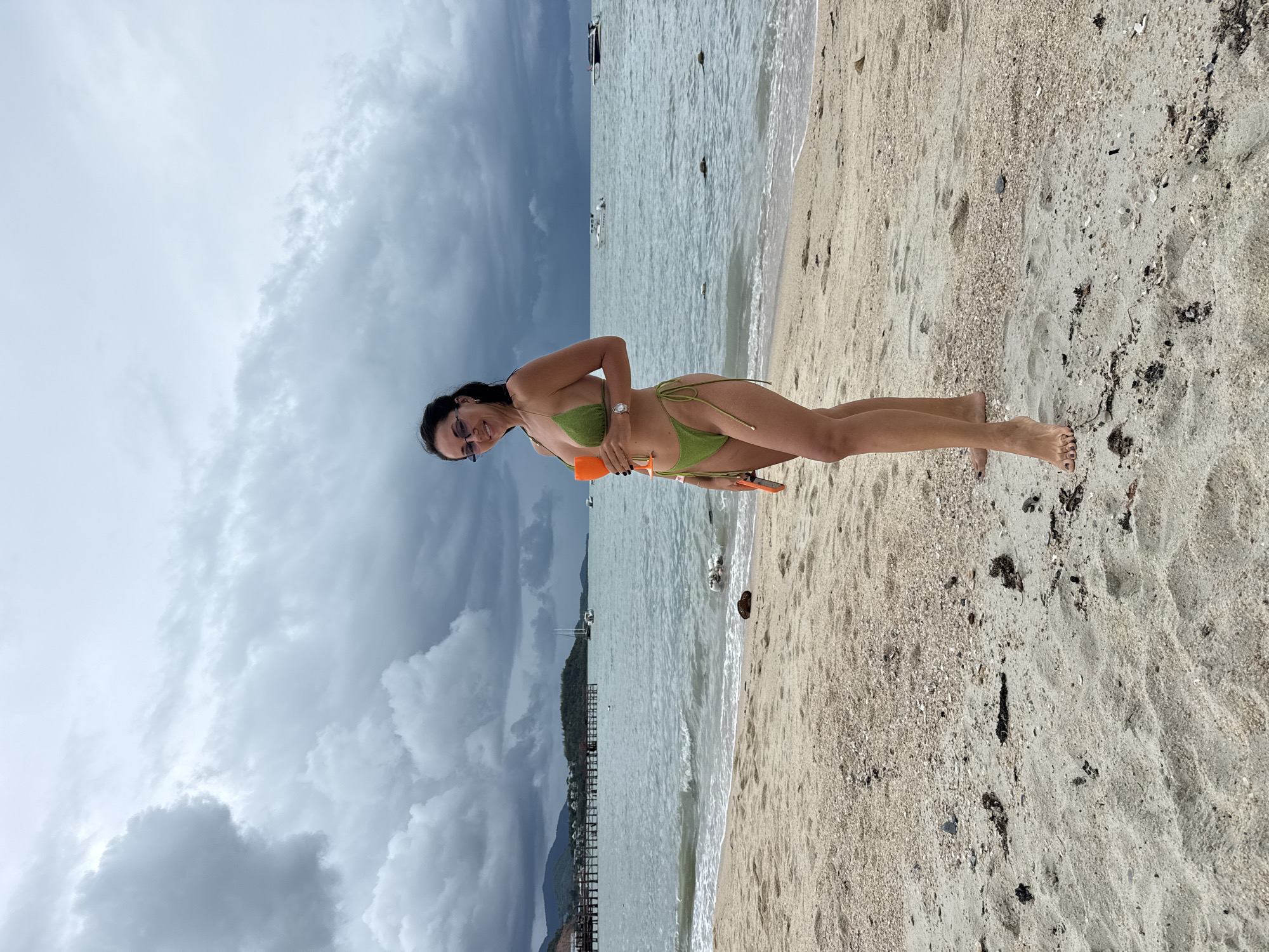 A woman stands on a sandy beach in the Ko Samui District of Thailand. She is wearing a green bikini and holding an orange object in her right hand. The ocean is behind her, with waves lapping at the shore. In the background, there are boats and a pier. The sky is cloudy, with dark storm clouds gathering. The sand on the beach is light brown and littered with small shells and debris. The woman is looking at the camera and smiling. She has dark hair and is wearing sunglasses. The overall impression is that of a person enjoying a day at the beach, despite the approaching storm.