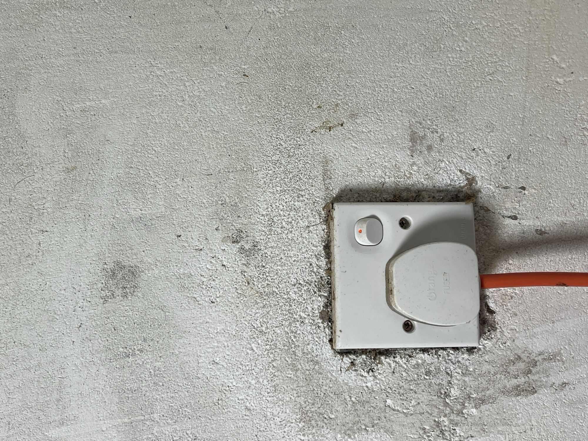 The image presents a close-up view of a white electrical socket and plug mounted on a textured, off-white wall. The wall, possibly concrete or stucco, exhibits a rough surface with visible dirt, discolouration, and possible signs of dampness or mold, especially around the base of the socket.
The electrical socket is a standard rectangular unit made of white plastic. It features a rocker switch on its left side, which is illuminated with a small, circular red light, indicating that the power is currently on. A white, three-pin electrical plug is inserted into the socket. An orange electrical cable extends from the right side of this plug.
Visible text on the plug includes a partially obscured brand name "Orange" and the word "FUSED", which is common for Type G plugs used in certain regions. Faint, indecipherable text is also present on the socket's faceplate above the plug. The screws securing the socket's faceplate to the wall are clearly visible.
The setting appears to be an interior space, likely a functional area such as a utility room, garage, or an industrial environment, due to the utilitarian nature of the electrical components and the unadorned, somewhat grimy appearance of the wall. No people or specific activities are depicted beyond the active electrical connection. The time of day is not discernible.