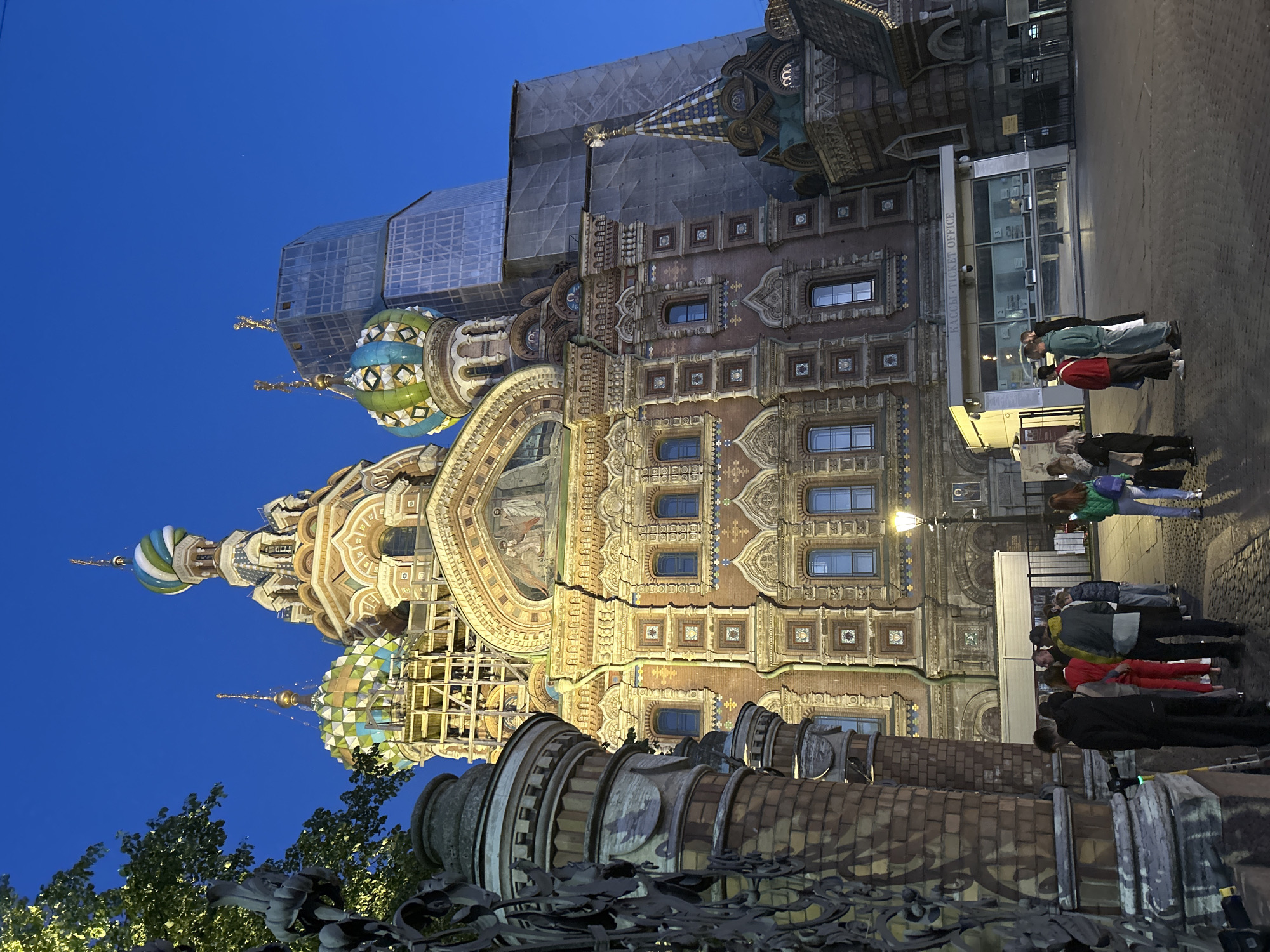 A low-angle, wide shot captures the Church of the Savior on Spilled Blood in St. Petersburg, Russia, at dusk. The ornate, red-brick cathedral with its distinctive onion domes, brightly colored mosaic tiles, and intricate architectural details is illuminated by artificial lights. Scaffolding and protective sheeting are visible on parts of the building and a modern glass-fronted skyscraper looms behind it. In the foreground, a group of about a dozen people, dressed in casual clothing, stand on a cobblestone plaza. Some are facing the church, others are conversing. A lamppost with a yellow light stands to the right of the group, and a decorative metal fence with pillars partially frames the left side of the image. The sky is a deep twilight blue, with no visible celestial bodies. A ticket office with "КАССЫ" (Ticket Office) signage is present to the right of the church entrance.