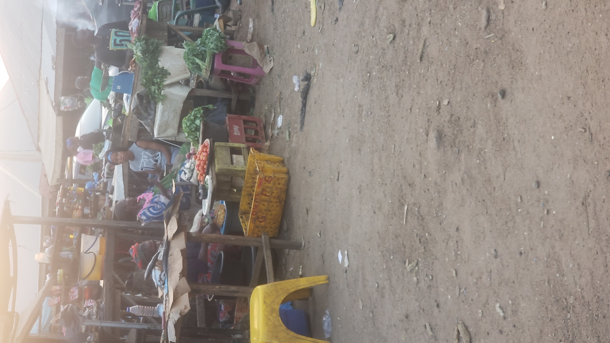 The image depicts a marketplace scene in Narayi, Nigeria. Several people are present, primarily vendors and shoppers engaging in commerce. Tables and makeshift stalls display various goods, including fresh produce like leafy greens and tomatoes. Recycled plastic crates and containers are used for storage and display. The ground is dirt, showing signs of foot traffic and litter. Overhead, a makeshift covering provides shade from the sun. The overall scene suggests a busy, informal economic activity in a developing urban area.