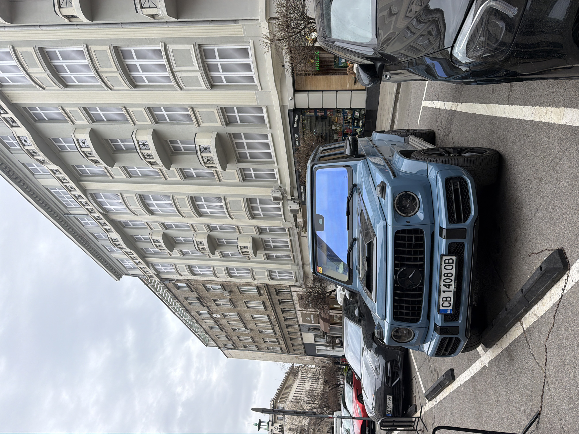 The image captures a street scene in Sofia, Bulgaria, featuring several parked cars, including a prominent blue Mercedes-Benz G-Class Brabus with license plate "CB 1408 OB". The vehicles are parked on a street marked with white parking lines and separated by black parking stops. Several other vehicles are visible, including a red vehicle and a black vehicle with license plate "CB 9541 HK".
In the background, a building with gray facade and symmetrical windows extends upwards, with "ROLEX" signage on the ground floor. Streetlights and pedestrian poles are also noticeable. The sky above is overcast.