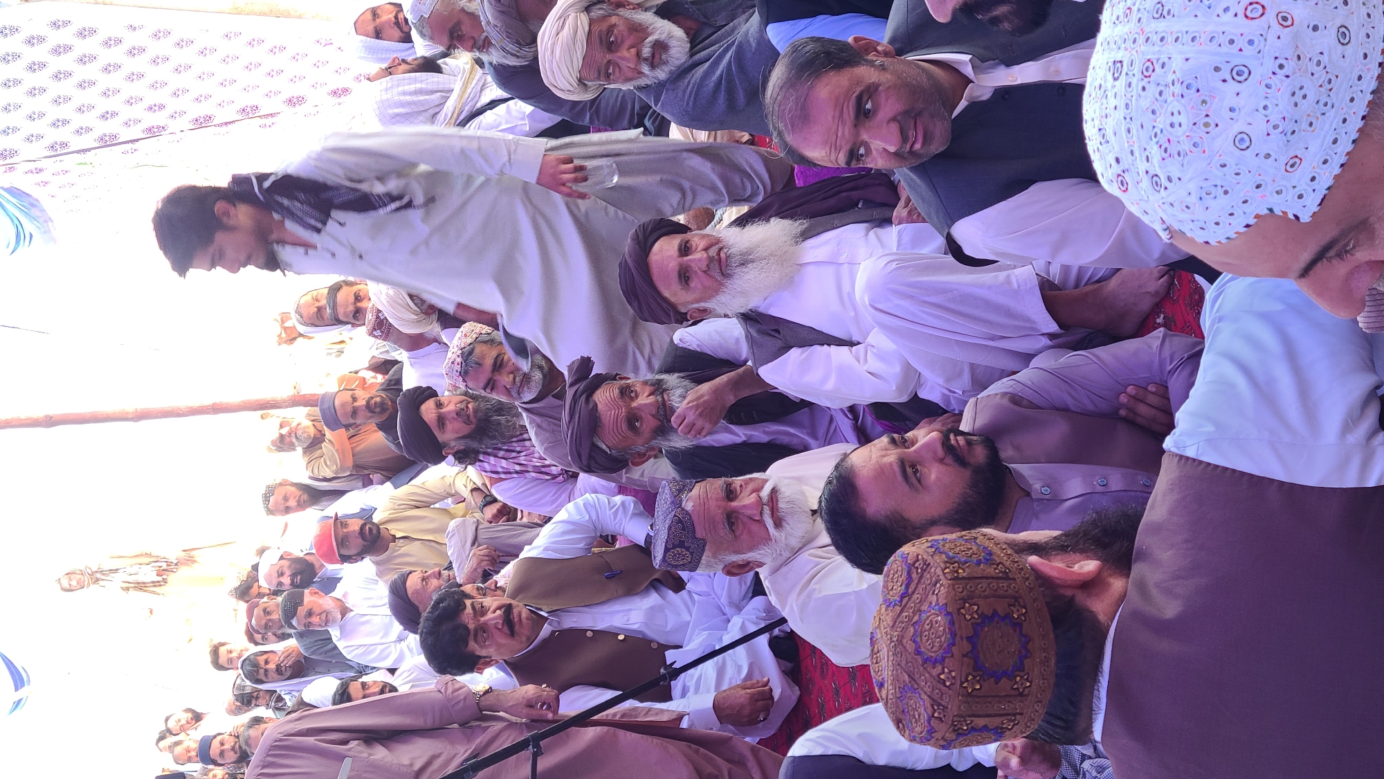 The image shows a gathering of approximately 30-40 men, likely in Panjpai, Pakistan. Most of the men are seated on the ground or on a low platform. They are dressed in traditional Pakistani clothing, including shalwar kameez, vests, and a variety of headwear such as turbans, skullcaps, and embroidered hats. Several men have beards, ranging from trimmed goatees to full, long white beards. Some are looking towards the camera, while others appear to be engaged in conversation or observing something outside the frame. The background is blurred, but a tent-like covering can be seen overhead with a simple floral pattern. A wooden pole is visible in the foreground. The overall impression is a community gathering or meeting.