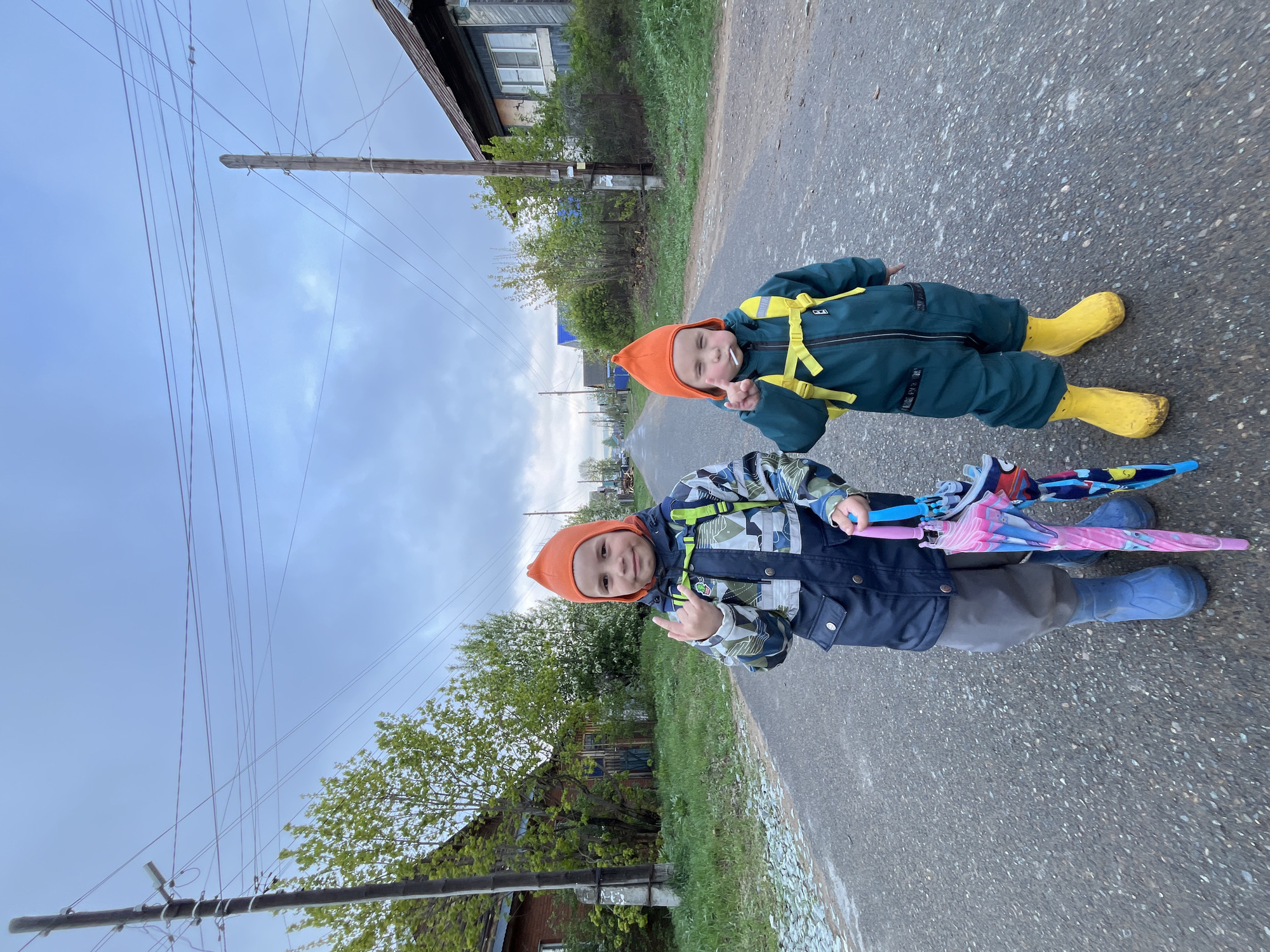 Two young children, both wearing orange hats and matching rain boots, are standing on a paved road. The child in the foreground is wearing a blue and camouflage jacket and holding two closed umbrellas. They are giving a thumbs-up gesture. The child behind them is wearing a green jumpsuit with yellow suspenders and holding up two fingers in a peace sign. The setting appears to be a rural or suburban street with residential houses and utility poles visible in the background. The location is Chaikovsky, Russia.