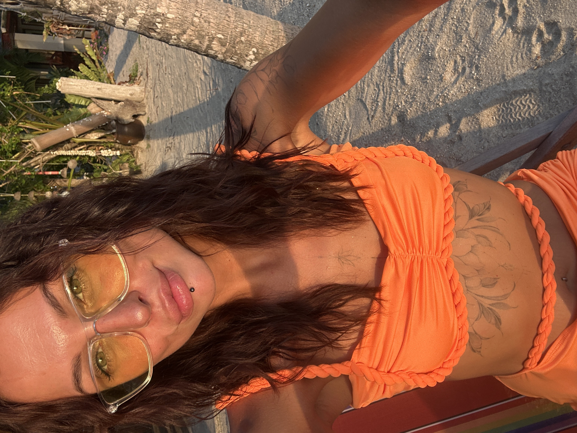 A woman with dark, wet hair and green eyes is seated on a striped lounge chair on a sandy beach, likely in Ko Samui District, Thailand, given the palm trees and tropical vegetation in the background. She is wearing a bright orange bikini with braided straps and trim. Her makeup is visible, including yellow-tinted sunglasses and a lip ring. She has tattoos on her chest and arm. The lighting suggests it is late afternoon or early evening, with warm, soft sunlight illuminating the scene. The beach appears relatively empty, with footprints visible in the sand.