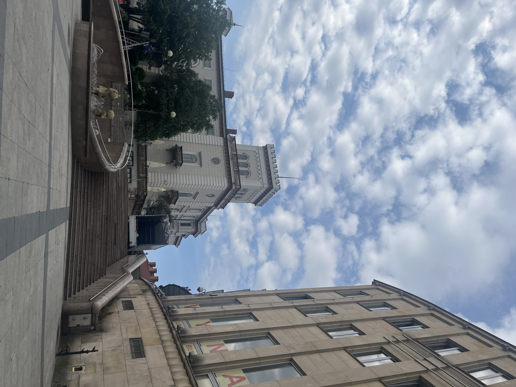 The image depicts a wide view of an urban square with a fountain in the foreground and buildings in the background. The square is paved with light-colored square tiles. A curved fountain with two small, gold-colored statues of children is visible in the lower left quadrant, with water spraying from it. To the right of the fountain, a wide, stepped staircase ascends towards the buildings.
The buildings are constructed with light-colored stone. The building on the left appears to be an older, ornate structure with a tower, possibly a castle or palace. It has arched windows and decorative elements. Trees with dark green foliage are planted in front of this building. The building on the right is a more modern structure with multiple stories and many windows. A large sign with red and yellow lettering is attached to its facade. A truck is parked in front of the building on the left, partially obscured by the stairs. The sky above is filled with white and gray clouds against a light blue background. The scene appears to be under daylight. There are no clear individuals captured in the act of interacting in the frame.