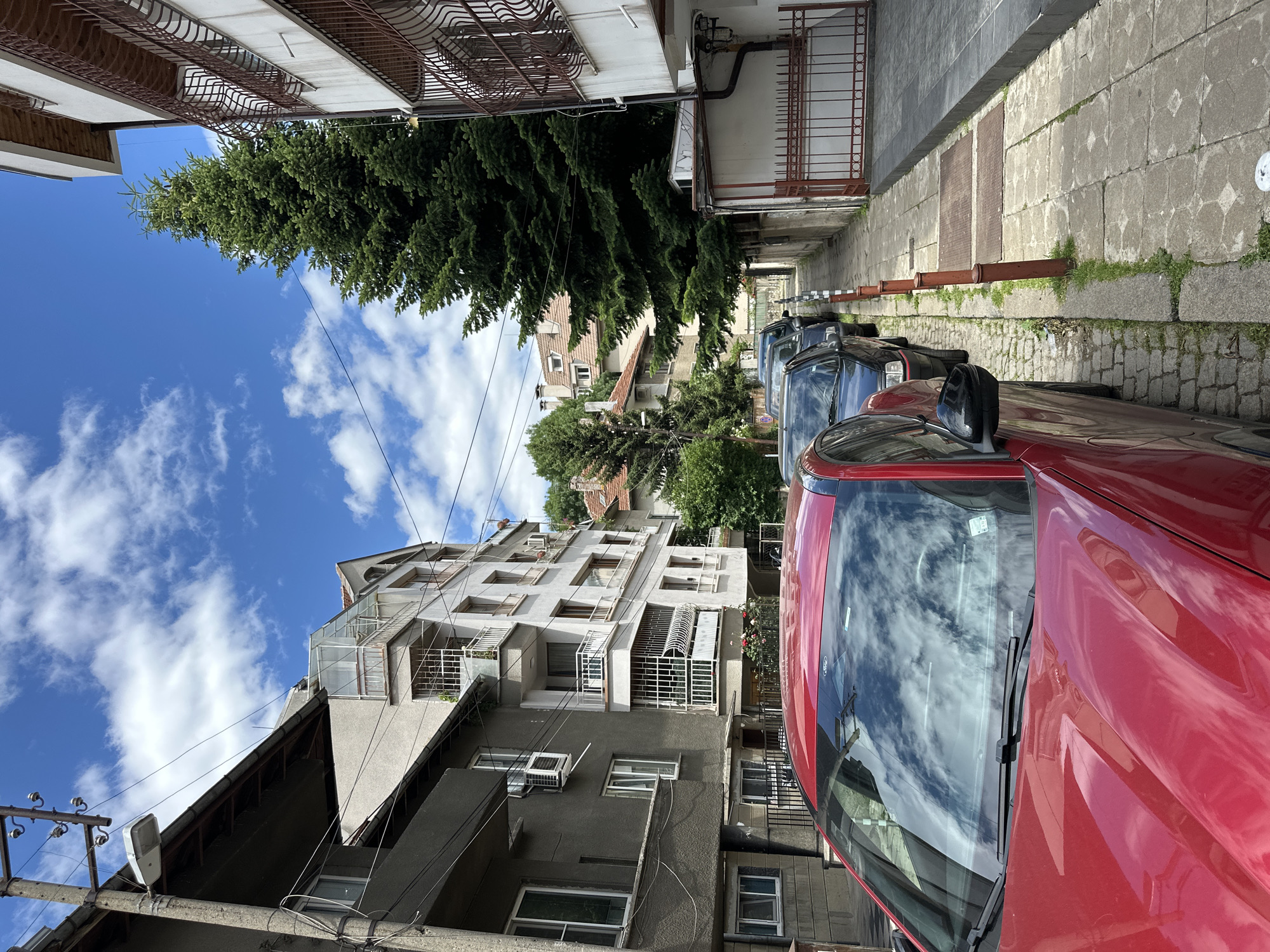 The image depicts a narrow street in Sofia, Bulgaria, lined with apartment buildings and parked cars. The buildings appear to be a mix of older, weathered structures and more modern ones, with balconies and windows visible. A tall evergreen tree stands prominently next to one of the buildings. The street itself is paved with cobblestones and tiles, with a sidewalk on the right. Several cars are parked along the street, including a red car in the foreground. The sky is blue with scattered white clouds. There are no people visible in the image. The scene conveys a sense of everyday urban life in a residential area.