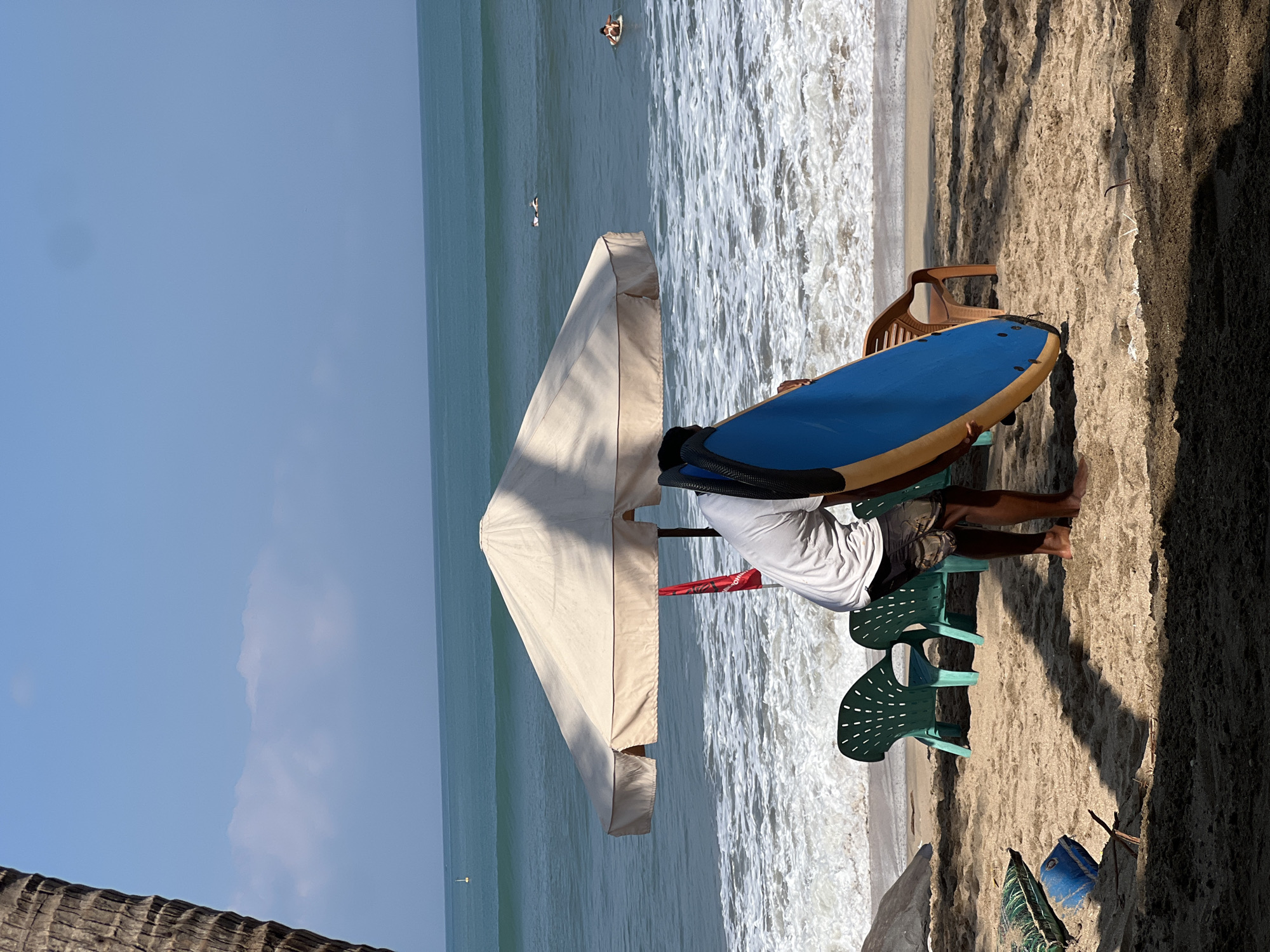 This image depicts a beach scene in Kuta, Indonesia, under a clear blue sky. In the foreground, a person is bent over, obscuring their face with a blue and yellow surfboard. They are wearing a white t-shirt and patterned shorts. Two green plastic chairs are positioned behind the person, near a large, light-colored beach umbrella. The sand is scattered with debris, including a blue plastic container and a green bag. The ocean is visible in the background, with waves breaking on the shore and several surfers in the water. A red flag is attached to a pole near the umbrella, and a palm tree trunk is partially visible on the left side of the frame. The scene is bathed in sunlight, casting shadows on the sand.