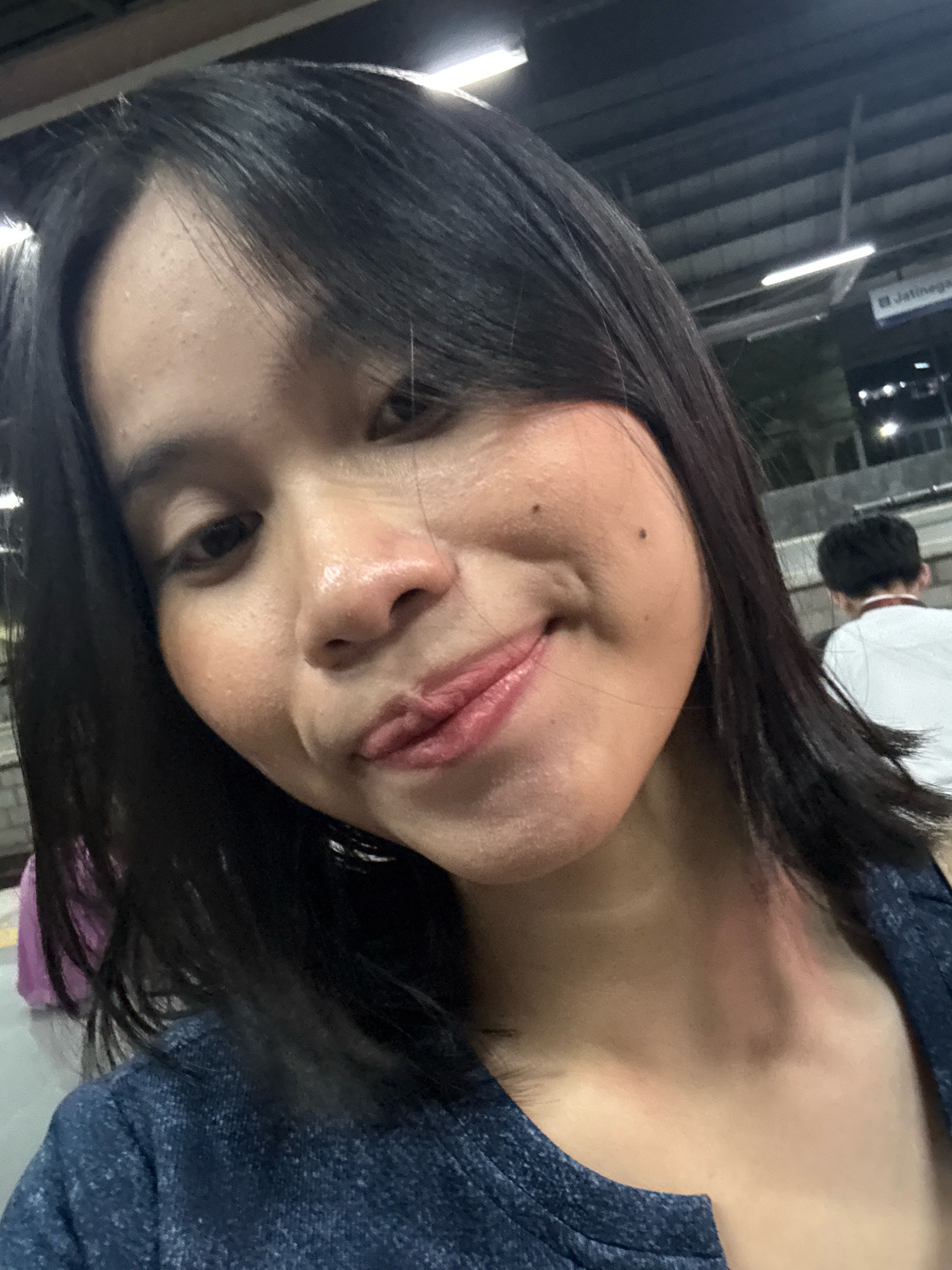 A young woman with dark hair and fair skin is taking a selfie. She is wearing a dark blue shirt and has a slight smile on her face. Her skin appears slightly dewy, and she has some faint moles on her cheeks. In the background, there is a sign that reads "Jatinegara," which is a district in East Jakarta, Indonesia. Other people are visible in the background, some out of focus, suggesting an urban environment, possibly a train station or a busy public area. The lighting suggests it is either evening or indoors with artificial lighting.