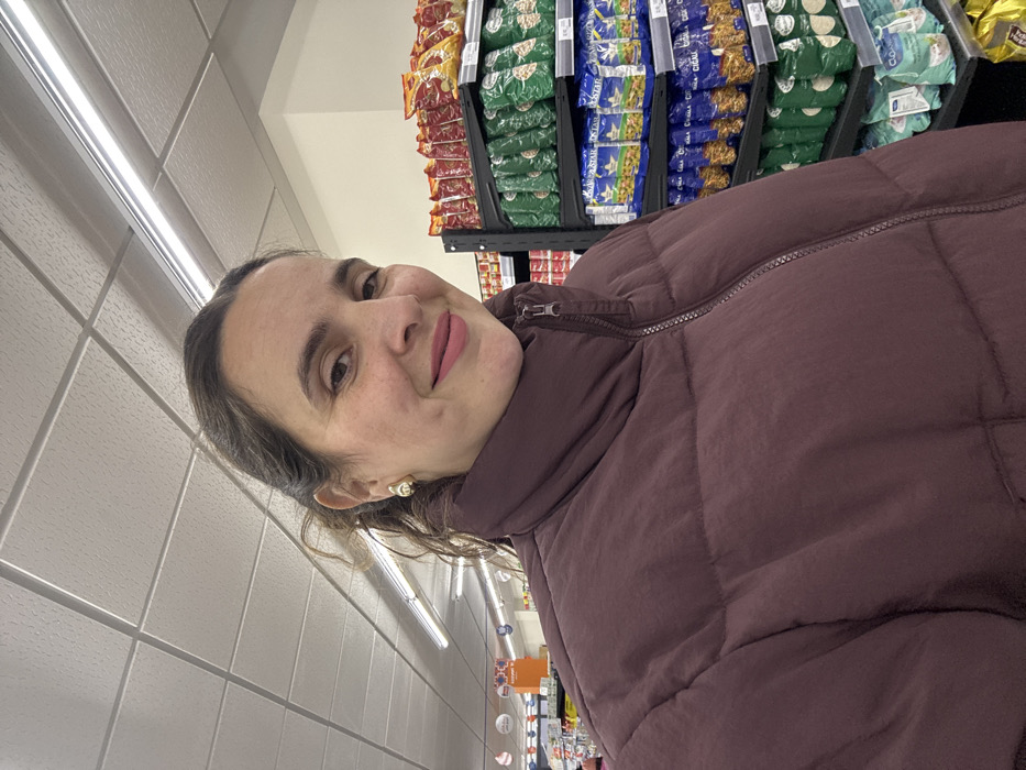 A woman with brown hair is in a store, wearing a maroon puffer jacket. She's looking directly at the camera and smiling. Behind her are shelves stocked with food items, including various packages of what appear to be snacks or pasta. The store has fluorescent lighting, and the walls are tiled. The photo appears to be taken from a slightly low angle.