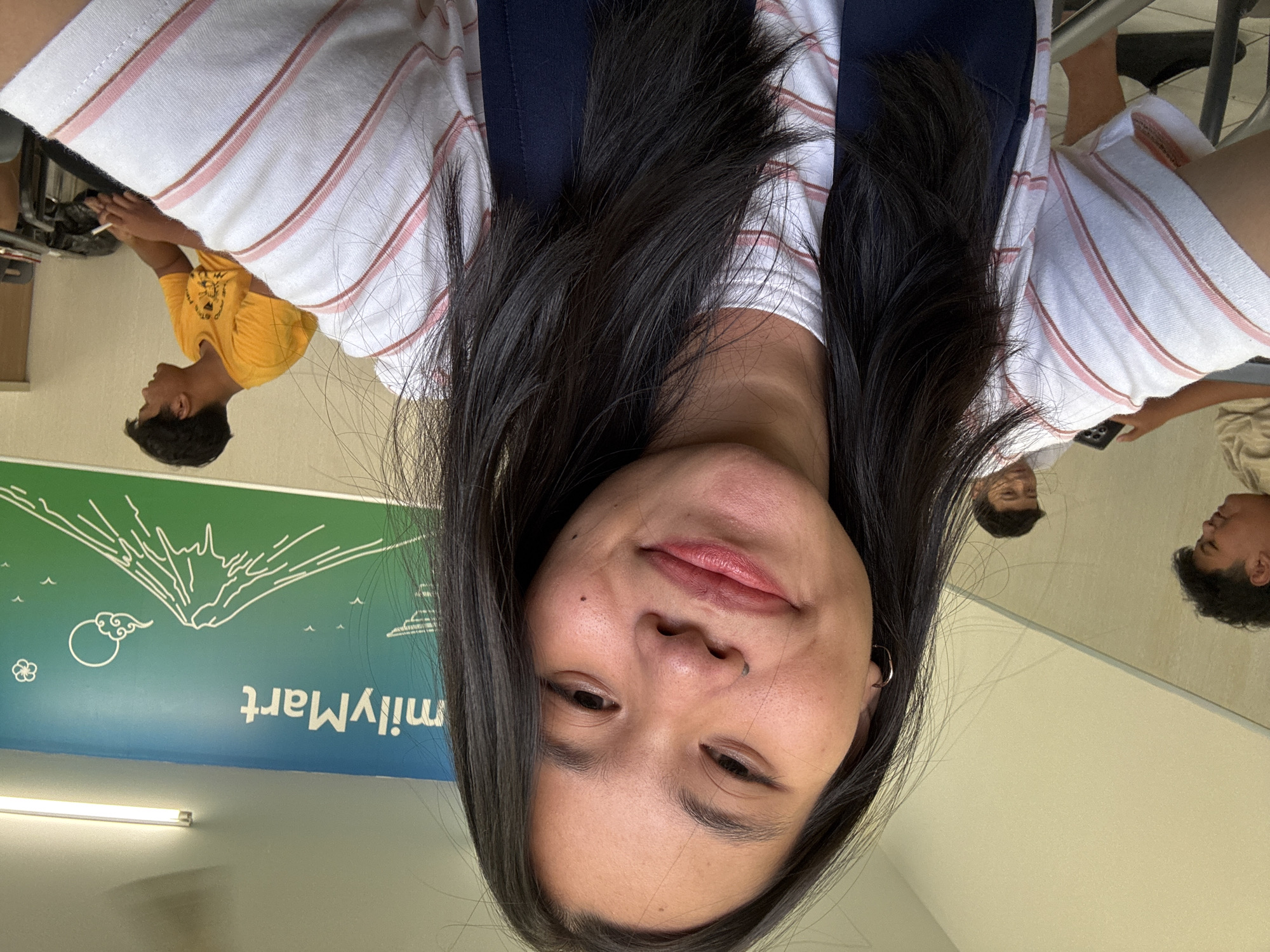 A young Asian woman with long, dark hair takes a selfie in a FamilyMart convenience store in East Jakarta, Indonesia. She is wearing a white t-shirt with pink stripes and a dark blue apron. Her face is tilted towards the camera, and she is smiling. In the background, a mural on the wall depicts Mount Fuji with the FamilyMart logo above it. Two other people are visible in the background: a young man in a yellow t-shirt and another person partially obscured. The lighting suggests the interior of a store.