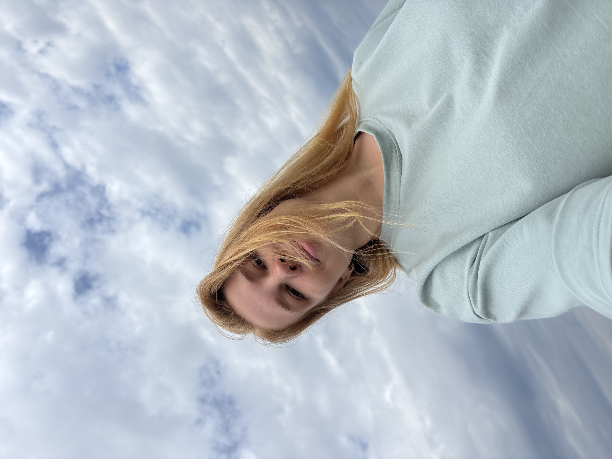 A low-angle, close-up selfie of a young Caucasian woman with shoulder-length blonde hair against a cloudy sky. She is wearing a light blue crew-neck sweatshirt. The image appears to be taken outdoors, possibly in the city of Breytovo, Russia, given the information provided. The sky is filled with dense, gray and white clouds, suggesting an overcast day. Her hair is slightly windswept, and she is looking towards the camera with a neutral expression.