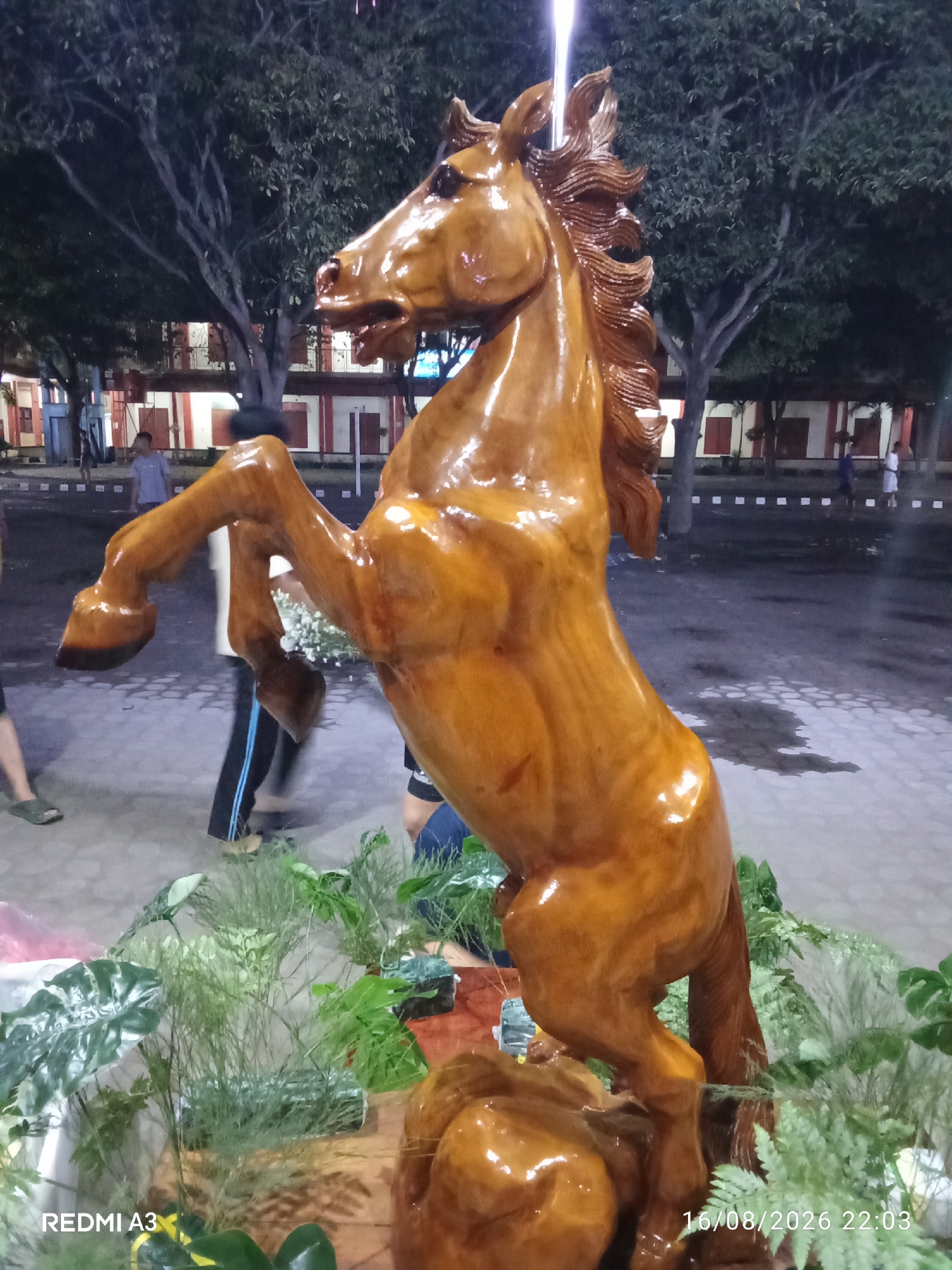The image shows a carved wooden sculpture of a horse rearing up on its hind legs. The sculpture is highly polished and has a warm, reddish-brown hue. It is surrounded by lush green foliage, including ferns and other leafy plants. In the background, there are buildings with red roofs and white walls, suggesting an outdoor setting in a built-up area. The sky is dark, indicating it is likely evening or night. There are a few blurred figures of people in the background, suggesting they are in a public space. The scene appears to be in the city of Surakarta, Indonesia, as indicated by the timestamp in the lower right corner.