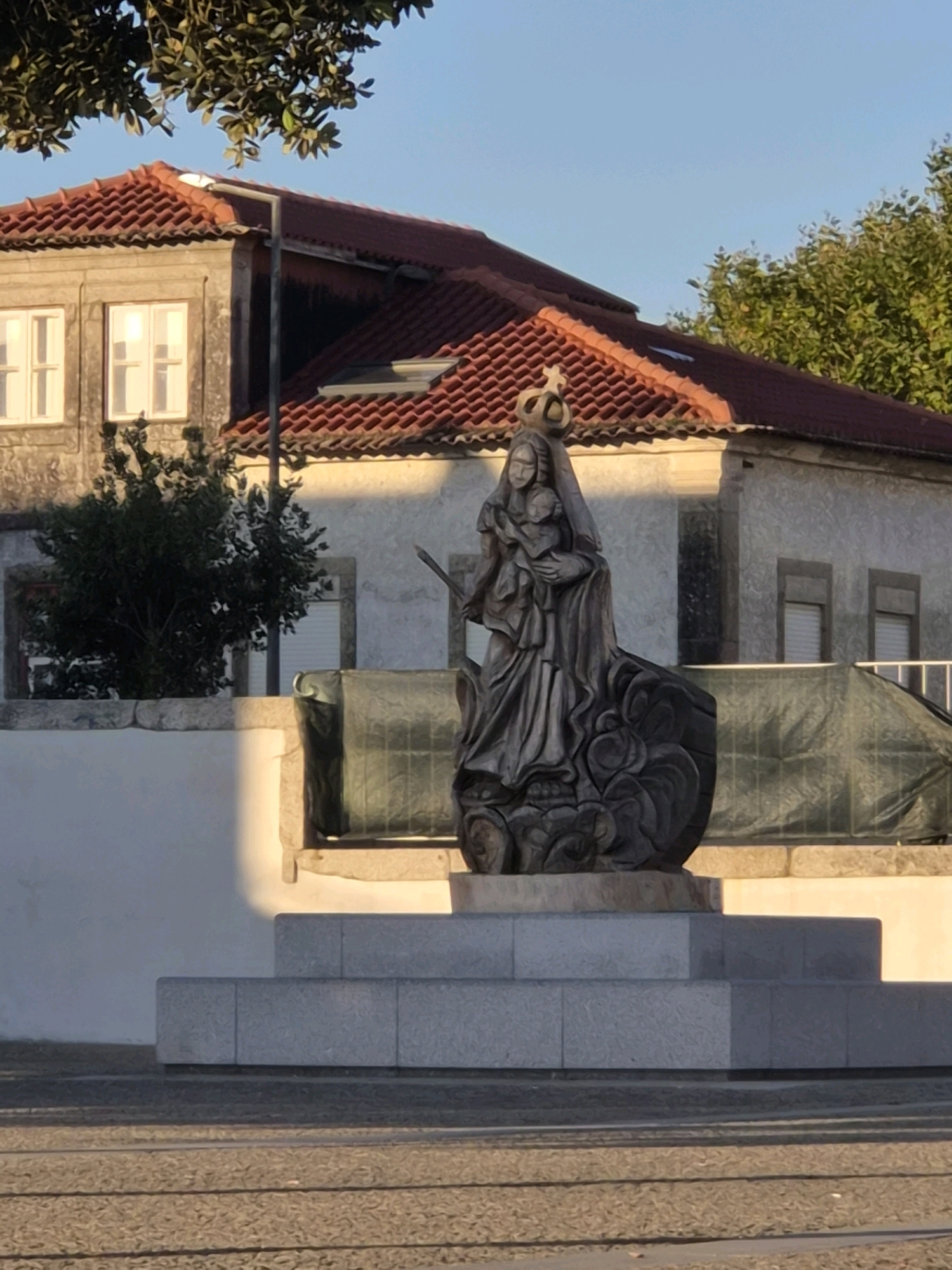 The image shows a stone statue of the Virgin Mary holding the infant Jesus, located in Esposende, Portugal. The statue is situated on a multi-tiered granite pedestal in front of a building with a red tile roof. The building appears to be residential or commercial, with windows and a balcony visible. Trees and foliage surround the statue and building, and the sky is clear and blue. The lighting suggests it is either early morning or late afternoon, with long shadows cast by the sun. The statue is dark in color and has a regal crown on its head. The base of the statue appears to be carved to resemble clouds or waves. The overall scene depicts a religious monument in an urban setting.