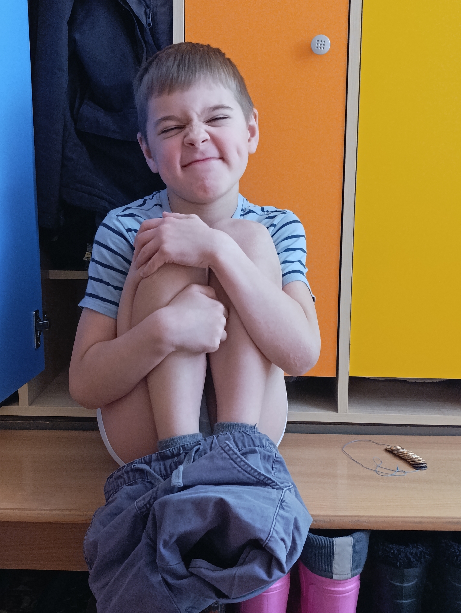 A young boy sits on a wooden bench in front of lockers. The lockers are blue, orange, and yellow. He wears a striped t-shirt and has gray shorts pulled down. He is hugging his knees and squinting with a grimace. A necklace and rain boots are visible. The location is Chaikovsky, Russia.