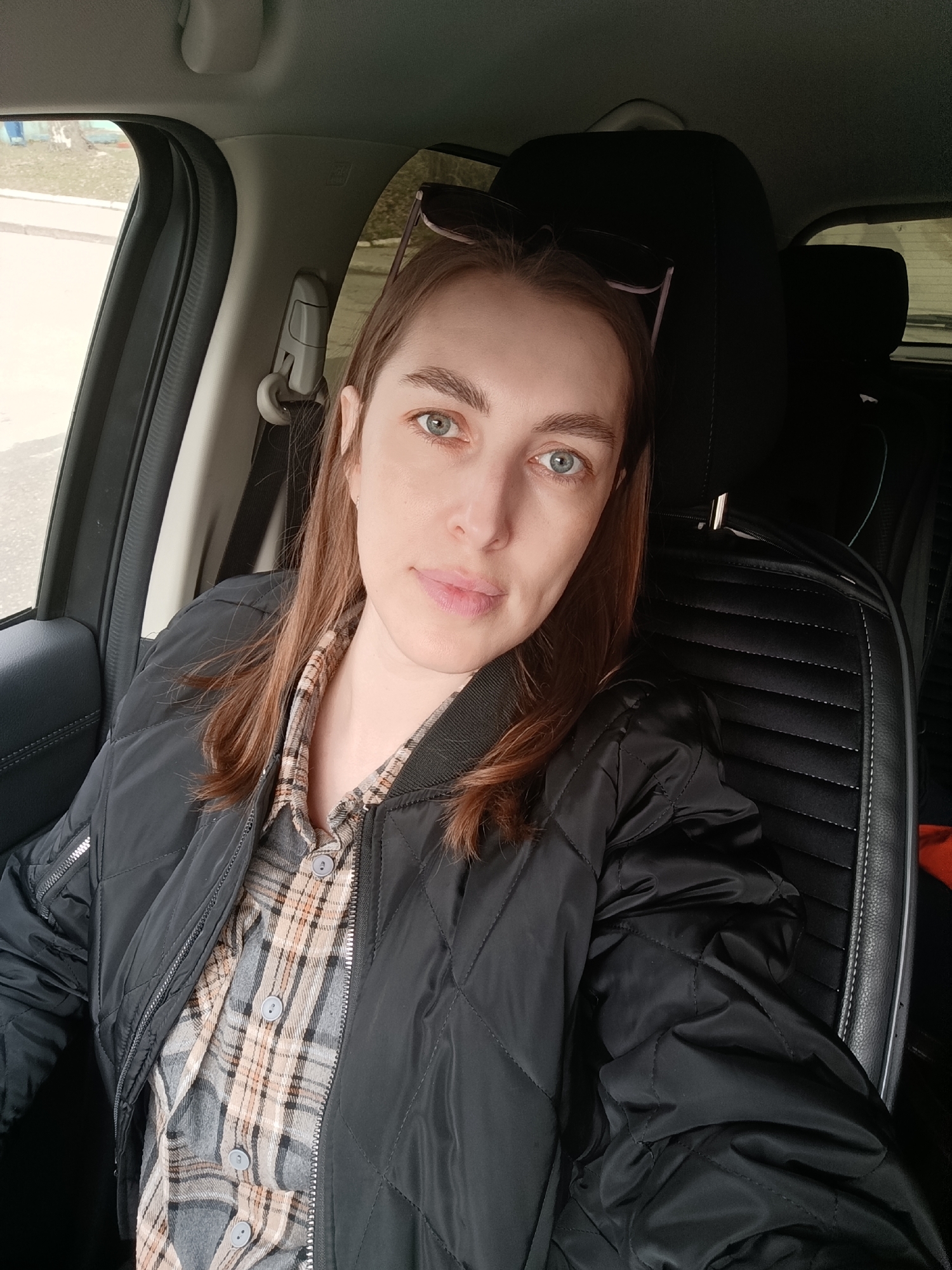 A woman is depicted sitting in the front seat of a car, looking directly at the camera. She has long, reddish-brown hair and light blue or blue-green eyes, with a neutral expression. She is wearing a black quilted bomber jacket over a plaid shirt in shades of grey, beige, and brown. A pair of dark-lensed sunglasses is perched on her head.
The setting is the interior of a vehicle. The car seat, a black, possibly leather or faux-leather material with horizontal quilted stitching, is prominently visible. A black headrest is behind her. Through the side window on the left, an outdoor scene can be seen, featuring a light grey or concrete surface, possibly a road or sidewalk, alongside a grassy area with some sparse, bare trees or bushes. The ambient light suggests it is daytime, possibly overcast, given the soft, diffused lighting.
The woman appears to be posing for a photograph, likely a selfie. Notable details within the car include parts of the ceiling, the door frame, and the B-pillar. On the B-pillar, a white label with the word "AIRBAG" is clearly visible. The overall impression is a casual moment captured in Chaikovsky, Russia.