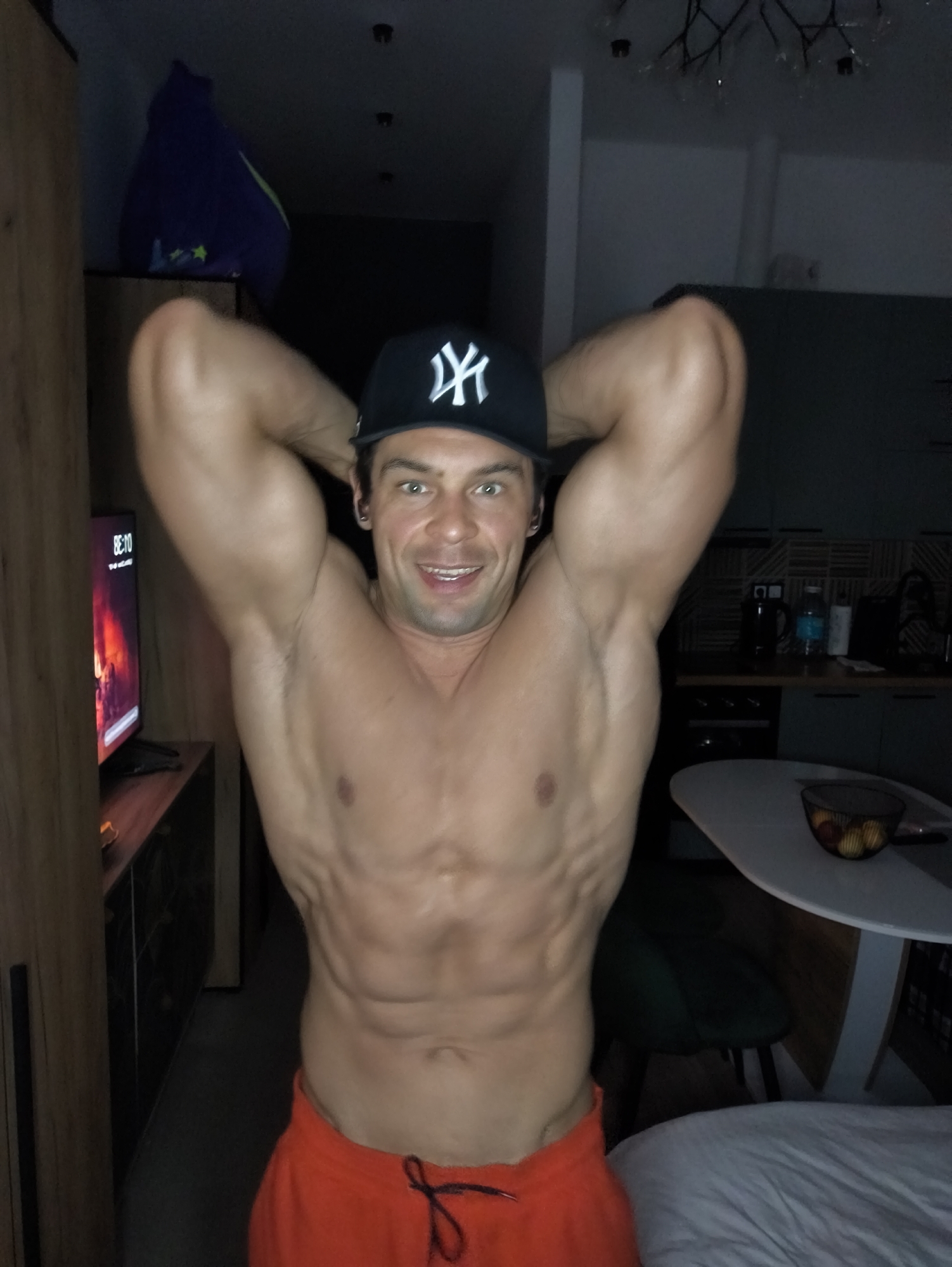 A muscular man wearing a black baseball cap with the New York Yankees logo and orange athletic shorts is posing for a photograph in what appears to be an apartment. He is shirtless and has his arms raised behind his head, showcasing his physique. The location is likely Moscow, Russia.
Behind him, to the left, is a wooden cabinet with a television displaying a scene with red and orange tones. To the right, there is a white round table with a bowl of fruit on it, along with a green chair. Further in the background, a kitchen area is visible with dark cabinetry and various appliances. The lighting in the apartment is dim, with overhead lights providing some illumination.