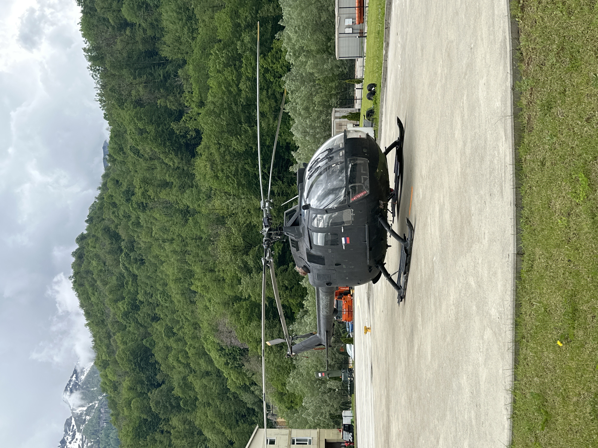 A black helicopter is parked on a concrete pad in Estosadok, Russia. Mountains covered in lush green vegetation are visible in the background, with snow-capped peaks partially obscured by clouds. A building and several vehicles are also visible in the background. The helicopter is oriented vertically, with its tail facing towards the right edge of the frame. Grass is on the right side of the concrete pad.