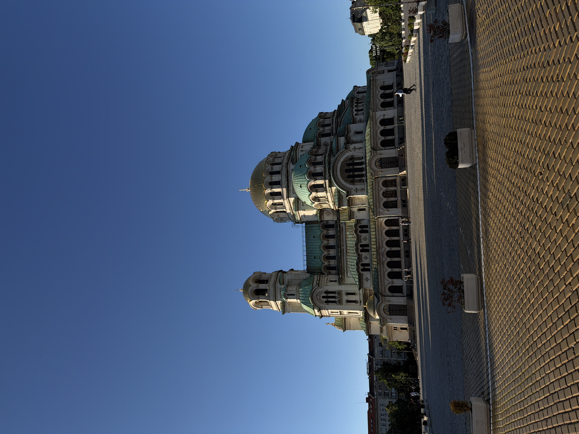 The image shows the Alexander Nevsky Cathedral in Sofia, Bulgaria, taken from a ground-level perspective. The cathedral's domes and architecture stand out against a clear, blue sky. To the right of the cathedral, a paved area is visible, made of small, yellow-brown bricks. There are also rectangular planters along the edge of the paved area, some containing small plants or bushes. In the background, there are buildings and trees, giving context to the urban environment. A person is also visible walking on the paved area.