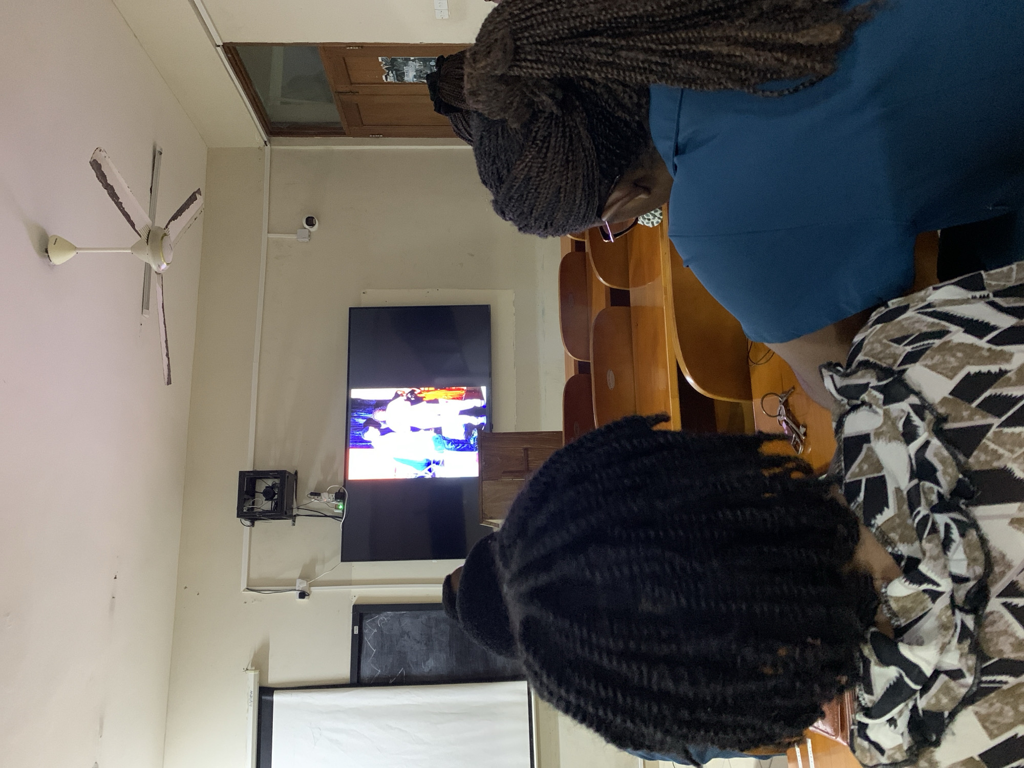 The image shows a room in Dar es-Salaam, Tanzania, with a ceiling fan, a security camera, and a television mounted on the wall displaying a sports event. Beneath the television is a small chalkboard and a projection screen. There are wooden benches arranged in rows, typically seen in a church or lecture hall. Three people are seated; two are facing towards the television and their hair braids are visible. One person is wearing a blue top and the other is in a black-and-white patterned top.