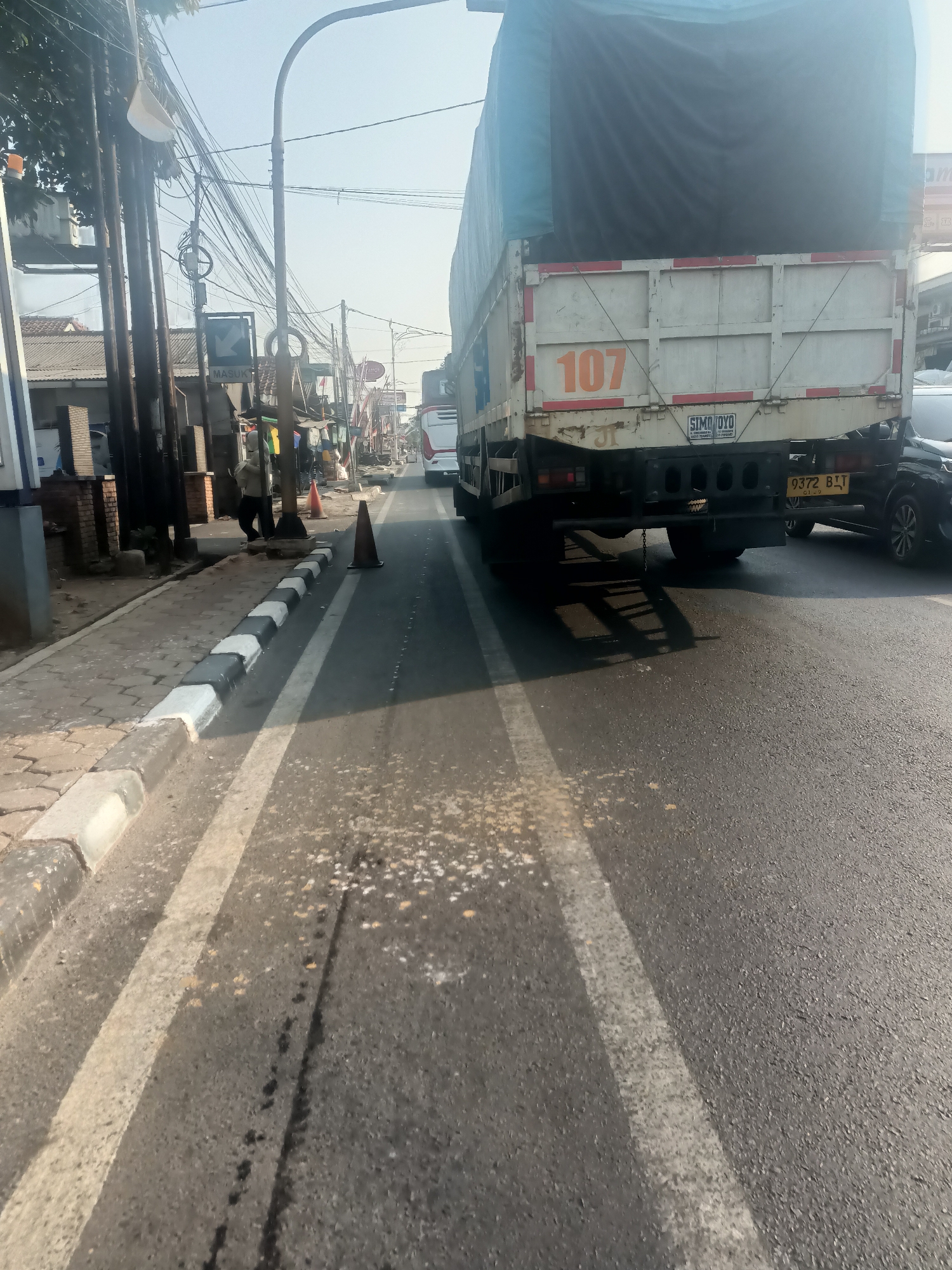 The image depicts a street scene in Purwakarta, Indonesia. In the foreground, the asphalt road is marked with white lines, indicating lanes. A truck with a blue tarp covering its cargo is prominent on the right side of the frame. The rear of the truck displays the number "107" and a license plate that reads "9372 BJJ". To the left, a sidewalk is visible with a decorative curb painted black and white. Beyond the sidewalk, there are buildings, poles with numerous electrical wires, and trees. A traffic cone is positioned on the road, and another is visible further down the street. In the background, other vehicles, including a white bus, are present, and the sky is clear. The environment suggests a typical urban or semi-urban setting with active road traffic.