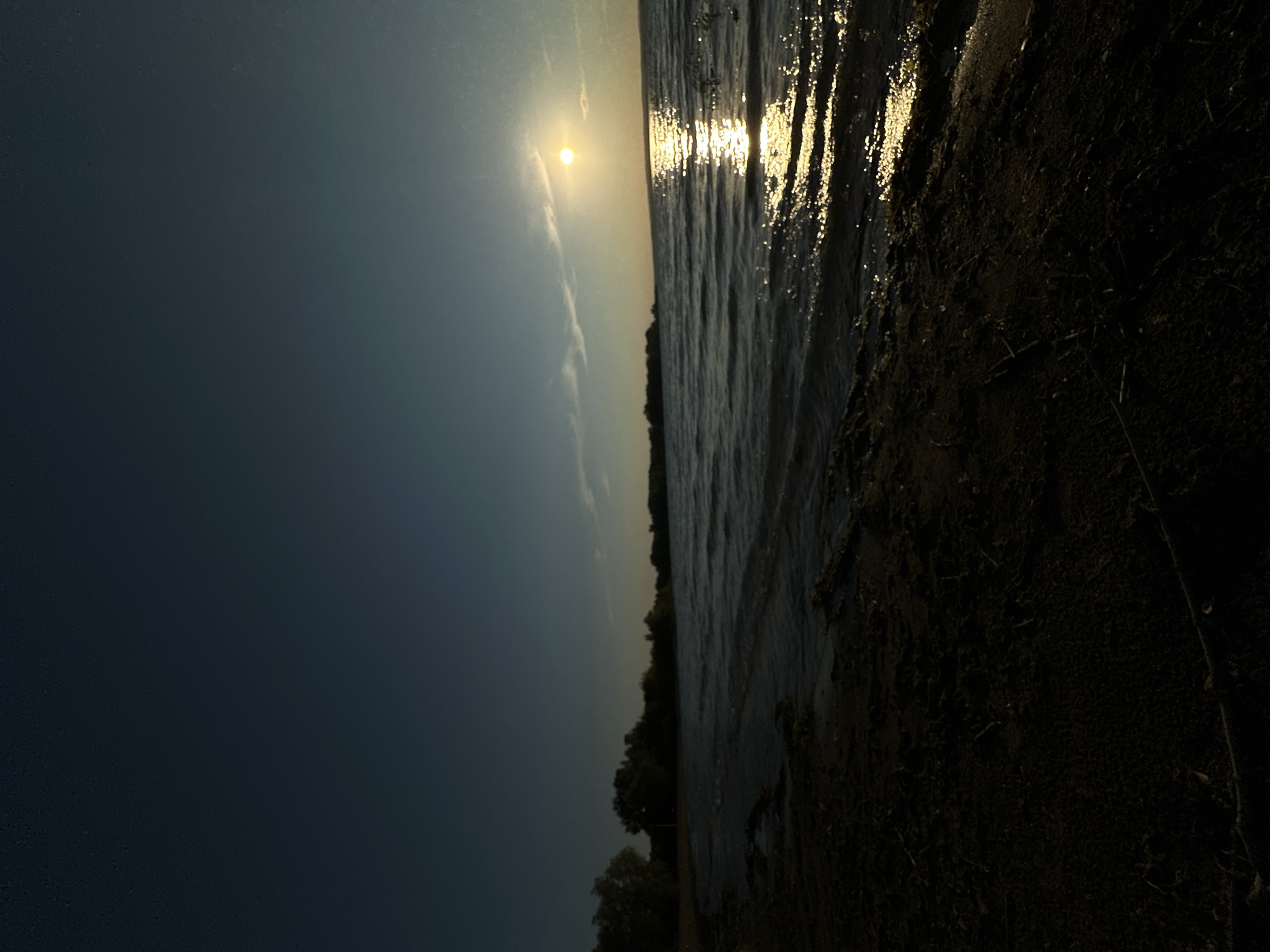 The image shows a body of water, likely a lake or sea, with a dark shoreline in the foreground. The sun is setting or rising, casting a bright reflection on the water. A distant treeline is visible on the opposite shore. The sky is a deep blue, with a few wispy clouds. The overall mood is serene and peaceful. The location is not specified.