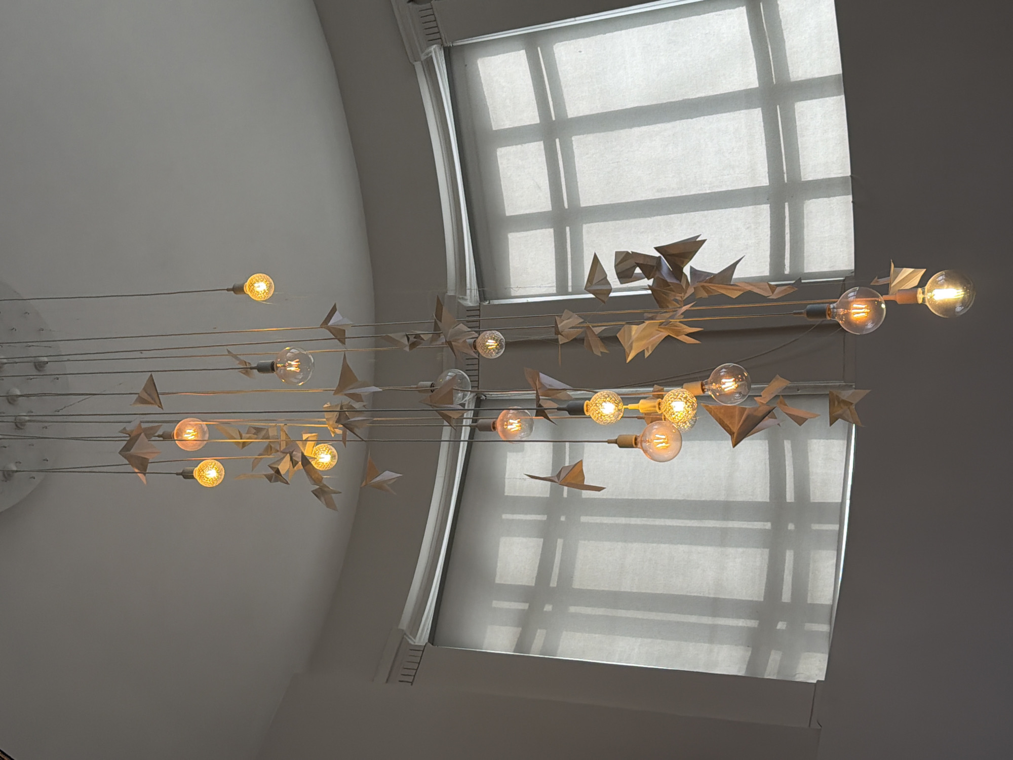 The image depicts an interior scene featuring a unique lighting fixture hanging in front of a window. The lighting fixture consists of several exposed light bulbs suspended on wires, each adorned with paper origami shapes resembling birds or airplanes. The window is large and features a white translucent shade that diffuses the light coming from outside. The surrounding walls and ceiling are painted in a neutral, light color, creating a minimalist and modern aesthetic. Based on the style and architectural details, the location is likely in a contemporary building or space. Without additional context, it's impossible to determine if this is Moscow, Russia, or not.