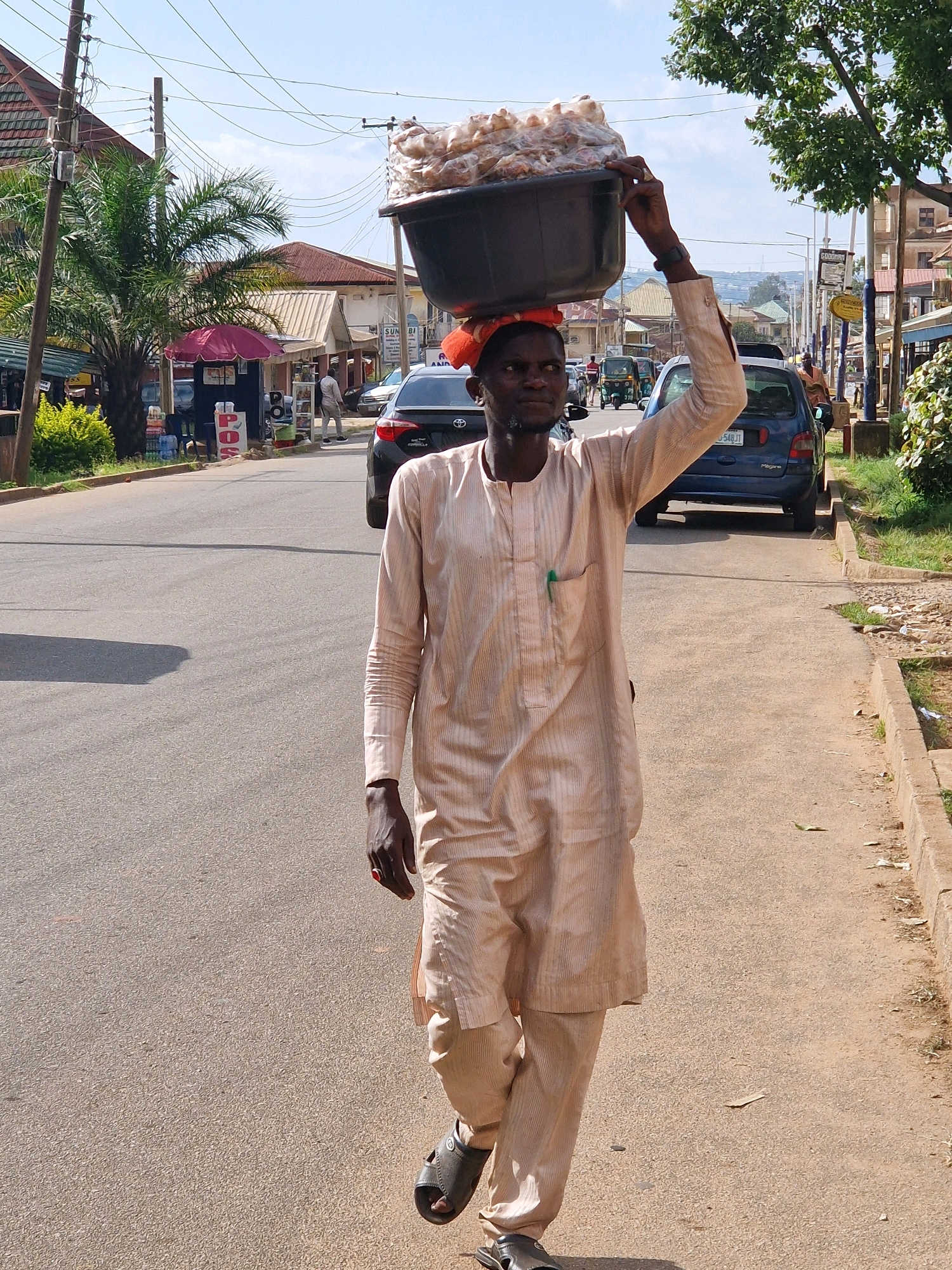 A man walks along a paved road in Jos, Nigeria, carrying a large black basin filled with food items on his head. He is wearing a light-colored, long-sleeved traditional outfit and sandals. The man is captured from the chest down, with his right hand supporting the basin. Behind him, cars are parked and driving on the road, and buildings line the street. Green trees and power lines are visible in the background, under a clear blue sky.
