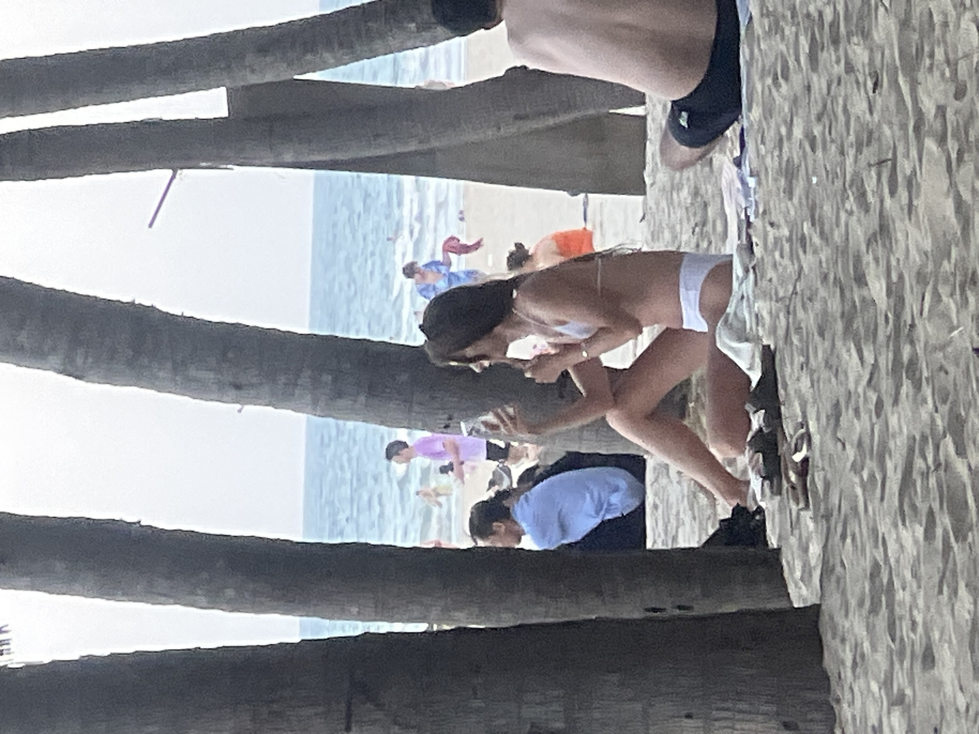The image depicts a beach scene in Da Nang, Vietnam, characterized by palm trees in the foreground and the ocean in the background. Several people are visible on the sand and in the water. A woman in a white bikini is sitting on a towel, holding a phone. Another person, partially visible from the back and wearing dark swim trunks, is also sitting on the sand. In the middle ground, a person in a light blue shirt is seated, and further back, people are seen walking and swimming in the ocean. The beach is sandy, and the water appears calm. The overall atmosphere is relaxed, suggesting a leisure activity on a warm day.