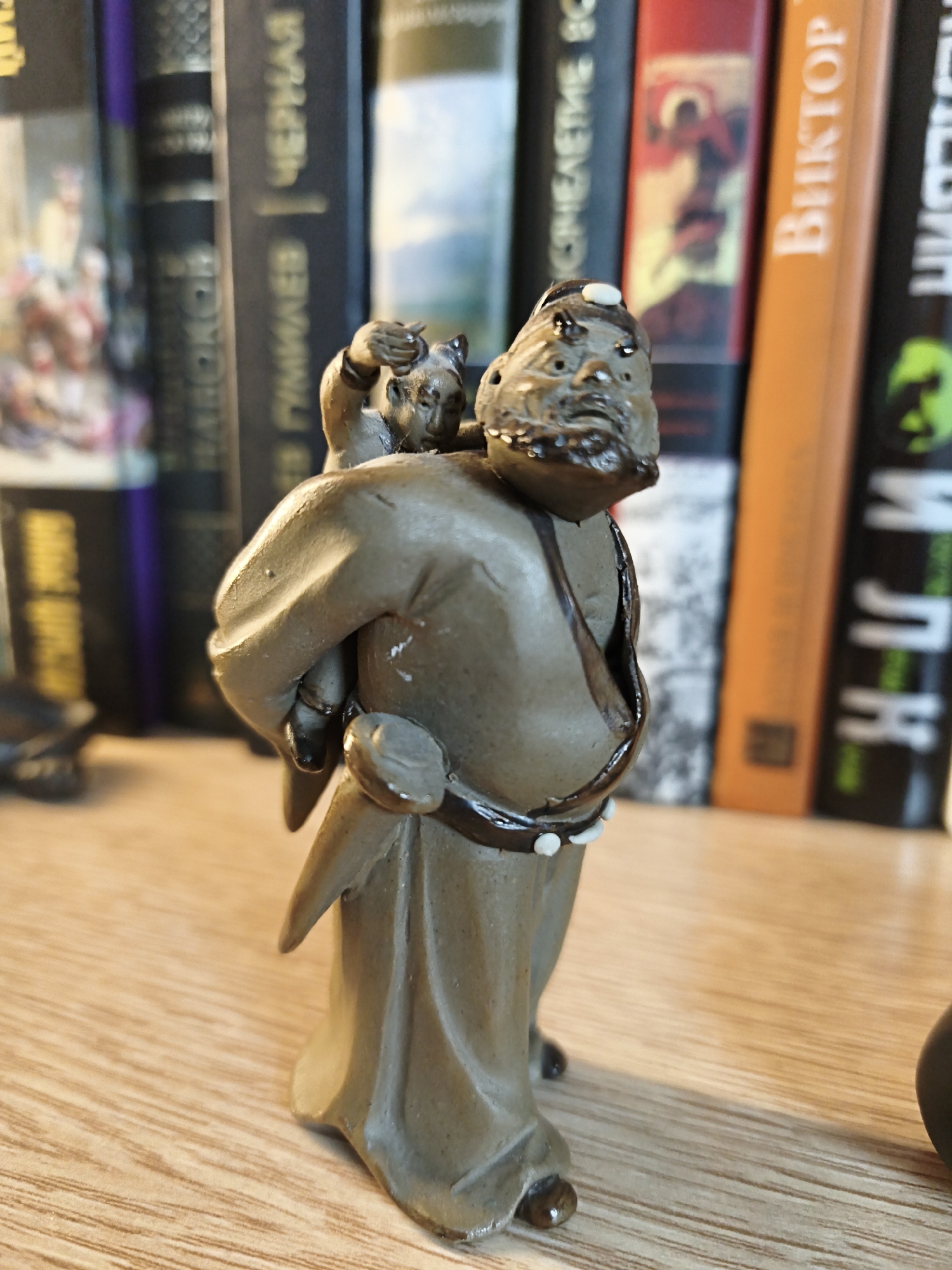 A small, figurine of a man carrying a child on his back stands on a wooden surface. The figurine is made of a material that appears to be clay or ceramic and is a muted brown color. The man has a beard and is wearing traditional clothing. The child on his back is smaller and appears to be reaching up to the man's head. Behind the figurine, a bookshelf filled with books is visible. Some of the book spines are in focus, showing Cyrillic text, while others are blurred. The overall setting appears to be indoors, possibly a home or a library. The lighting suggests it is daytime. The figurines are the central focus of the image, and the books provide context for the environment. The image is taken from a slightly low angle, emphasizing the figurines. There is no discernable action taking place other than the static pose of the figurines.