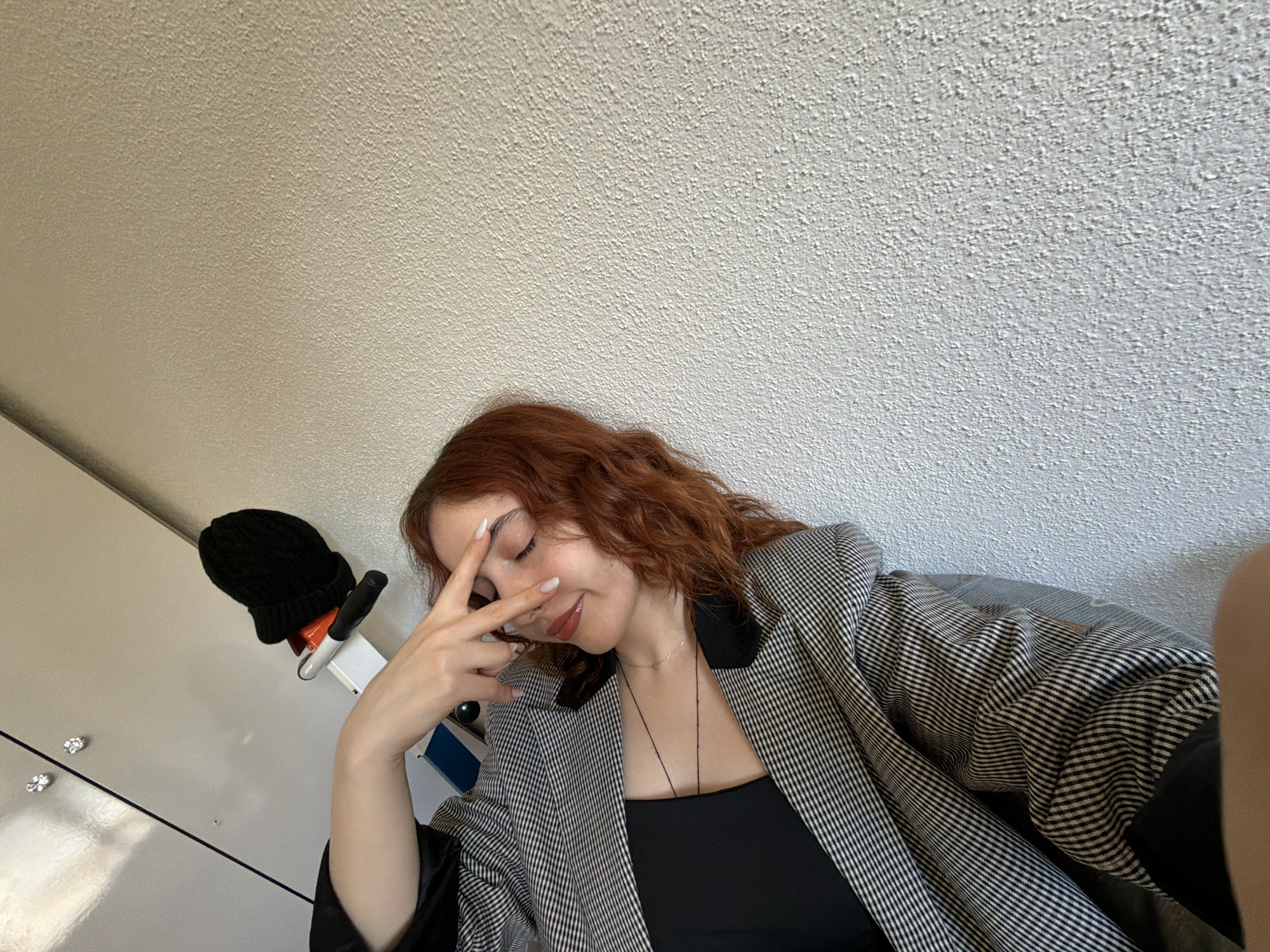 A woman with auburn, curly hair is posing indoors. She is wearing a black top underneath a houndstooth-patterned blazer. Her right hand is raised, fingers spread, partially obscuring her face. She is wearing a delicate necklace. To her left is a black knitted hat placed on a white shelf-like structure mounted on a white surface, likely a cabinet or door. The background consists of a textured, off-white ceiling. The location within Strasbourg, France, cannot be determined from the given image.