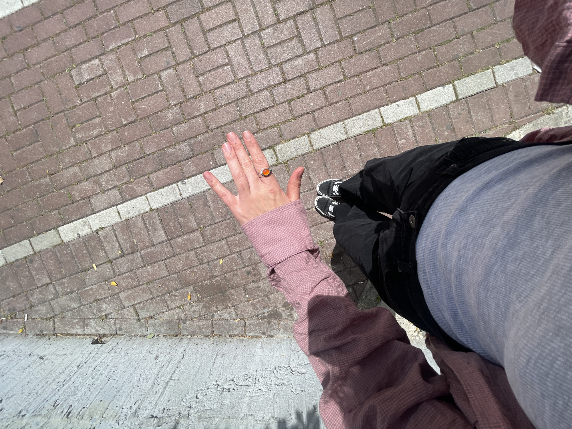 A person stands on a brick and concrete paved surface. They are wearing black jeans, a light blue t-shirt, and a pink checkered long-sleeved shirt. They have a ring with an orange stone on their left hand. The brick pavement has a white painted stripe. The setting appears to be outdoors, likely in an urban area with brick paving and concrete surfaces. While the location could be Rafailovići, Montenegro, there is no evidence within the image to confirm this.