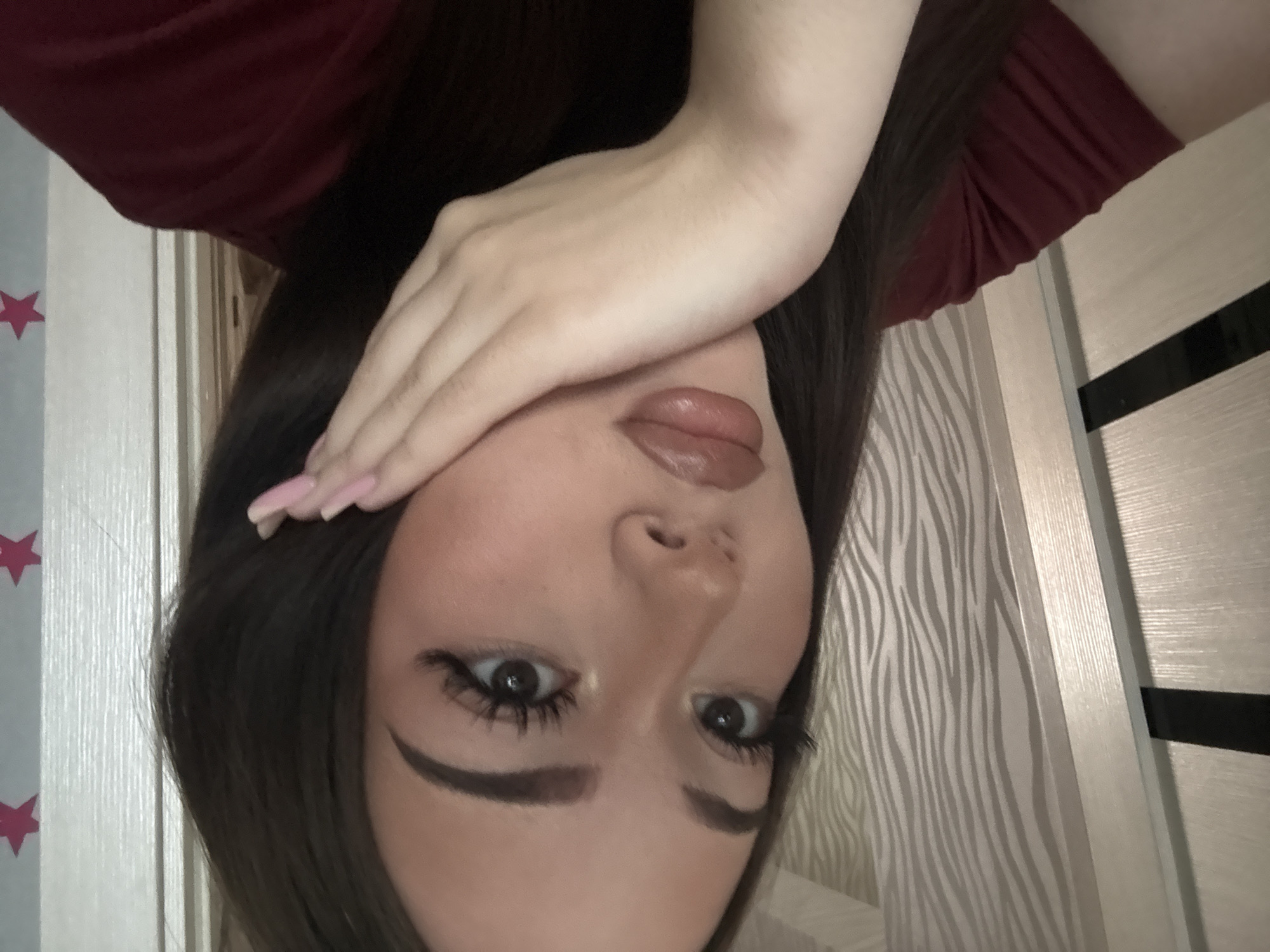 A close-up, upright portrait features a young woman indoors, presumably in Naberezhnye Chelny, Russia. She has long, dark hair and is wearing a dark red top. Her right hand, adorned with long, light pink painted nails, rests across her upper face, partially covering her forehead and one eye. Her visible eye is open and looking towards the camera, featuring prominent dark eyelashes and well-defined eyebrows. Her lips are slightly parted, showcasing a light, glossy color.
The background on her right (viewer's left) includes a light-colored wall decorated with small red star decals. To her left (viewer's right), a wall is covered with wallpaper featuring a distinct grey and white zebra-stripe pattern. A light-colored door frame and a light wooden door with a dark horizontal accent stripe are also visible in the background. The lighting suggests an indoor setting, likely daytime or well-lit by artificial light. The overall mood conveyed by her pose and gaze is one of a direct, perhaps alluring, connection with the viewer. No visible text is present in the image.