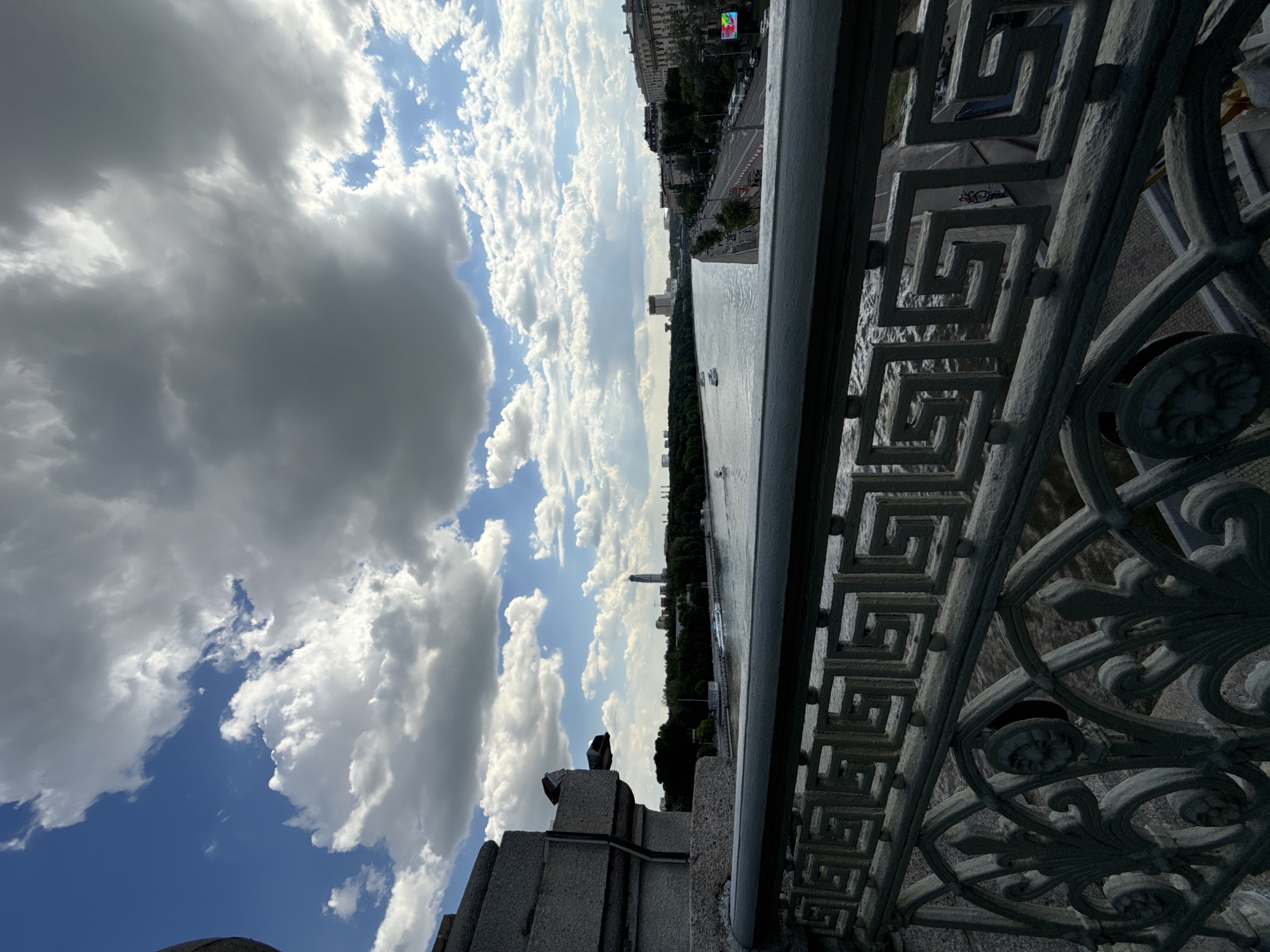 The image captures a panoramic view of a city, identified as Moscow, Russia, from an elevated vantage point, likely a bridge or embankment. An ornate, dark grey metal railing dominates the right foreground, featuring a distinct geometric meander pattern on its upper section and intricate scrollwork with circular medallions or faces below. Adjacent to the railing is a sturdy grey stone or concrete structure.
Beyond the foreground elements, a wide river stretches across the midground, its surface reflecting the daylight. Several boats or small ships are visible on the water, indicating active river traffic. The far bank of the river is lined with dense green trees, behind which urban architecture emerges, including a distant tall, slender building.
Further across the river, on the left side of the frame, lies a vibrant cityscape. A wide road with vehicles runs parallel to the river, flanked by multi-story buildings. A large, brightly lit digital billboard or screen displaying colorful, indecipherable content is noticeable on one of the buildings.
The sky occupies a significant portion of the image, filled with a dramatic interplay of large, fluffy white cumulus clouds and darker grey clouds, broken by patches of clear blue sky. The scene depicts a bright, partly cloudy day, suggesting either late morning or afternoon. No specific human activity is central, but the presence of vehicles and boats indicates an active urban environment. No decipherable text is visible.