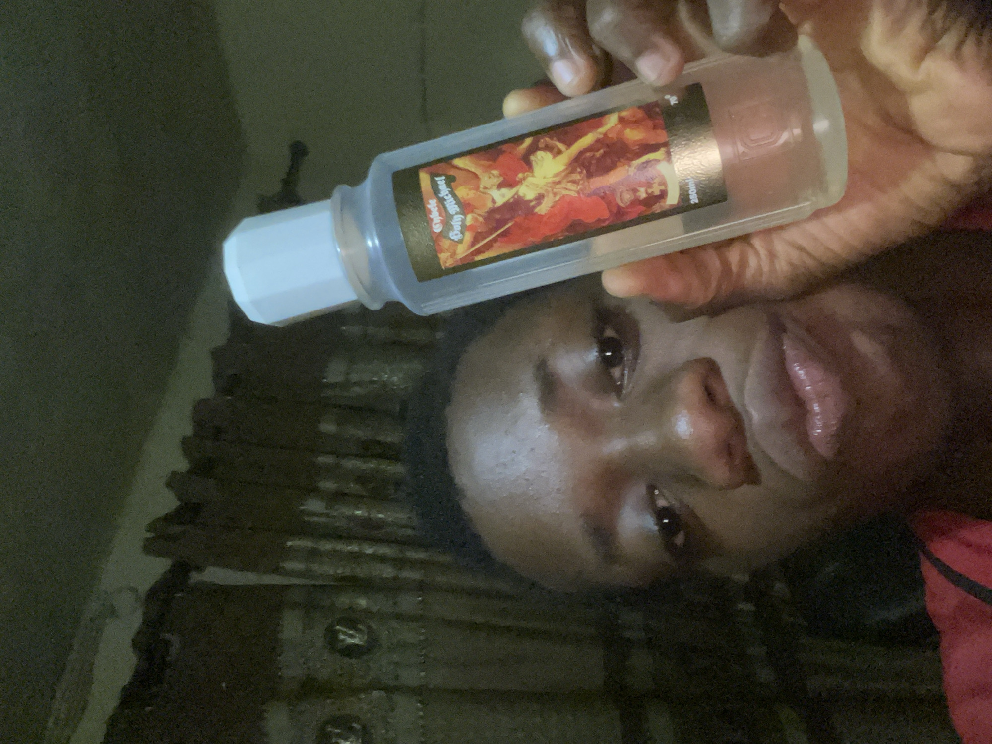 An image taken in Onitsha, Nigeria, features a person with dark skin holding a clear plastic bottle. The person's face is partially visible, looking towards the camera with a neutral expression. They are wearing a red garment, partially visible on the right side of the frame. The bottle has a white, hexagonal cap and contains a light-colored liquid.
A rectangular label is affixed to the bottle, displaying a dark background with a vibrant, classical-style painting of multiple figures rendered in red, orange, and yellow tones. Visible text on the label identifies the product as "Cybele Black Soap" and indicates a volume of "250ml". The person's hand is wrapped around the bottle, holding it up. The background is dark and indistinct, indicating an indoor setting with subdued lighting. The scene captures a casual moment, possibly a self-portrait or a product showcase.