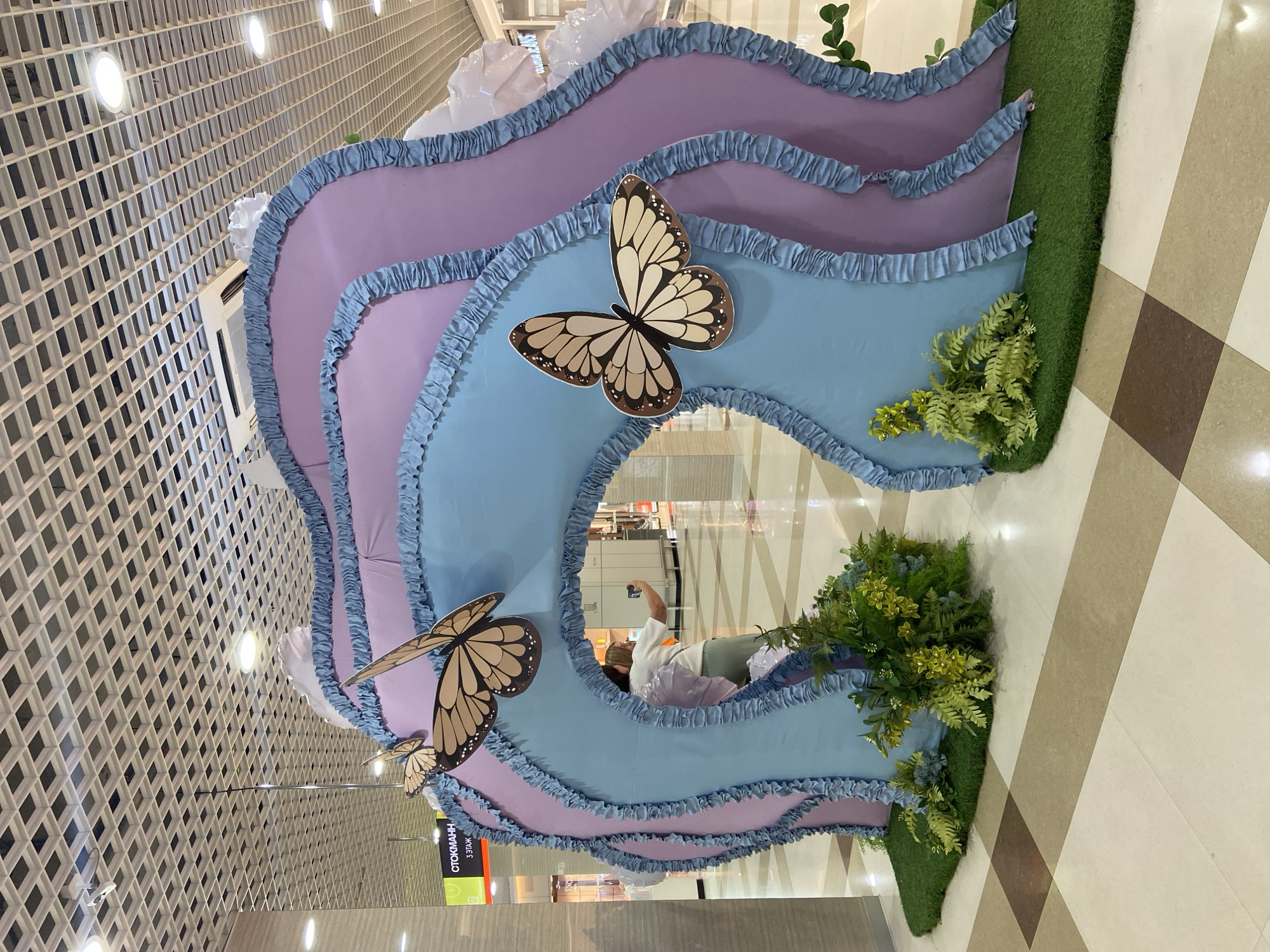 The image shows a decorative archway in a shopping mall. The archway is made of fabric in shades of blue and purple, with ruffled blue trim. Two large, brown and cream-colored butterfly decorations are attached to the arch. Artificial greenery and grass cover the base of the archway. In the background, a woman is taking a selfie in front of the archway. A sign in Russian that reads "СТОКМАНН 3 ЭТАЖ" (Stockmann 3rd Floor) is visible on the left. The ceiling of the mall is visible at the top of the image, with a grid-like structure and circular lights. The floor is tiled with large squares of varying neutral colors. The scene is likely located in Irkutsk, Russia, given the Russian text on the sign.