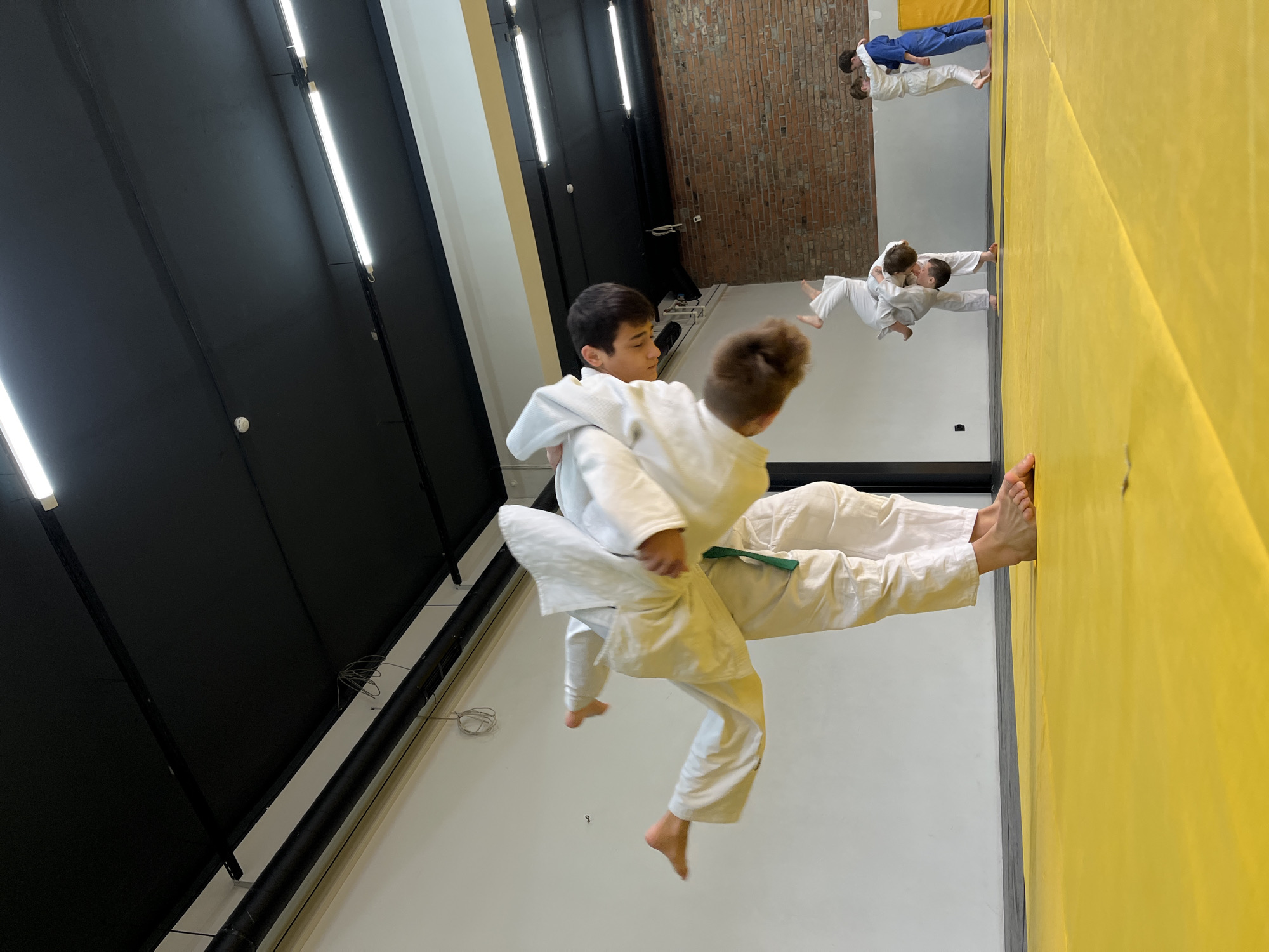 Two young judoka wrestlers are grappling in a dojo. One wears a green belt. A mirrored wall shows other judo participants. The background includes a brick wall and bright lights. The location appears to be Krasnoyarsk, Russia.