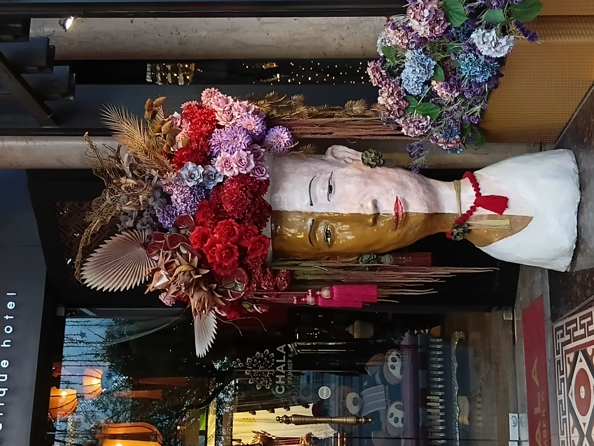 An ornate, head-shaped sculpture with a split gold and white face is adorned with a large, vibrant floral arrangement on its head, featuring red, purple, and pink flowers, as well as dried foliage. The sculpture is positioned in front of a building entrance, with its left side partially obscured by a glass door. Reflected in the glass are orange lanterns, trees, and a sign that reads "CHALA NUMBER 6". To the right of the sculpture, a basket overflowing with purple and blue hydrangeas is visible. The sculpture is wearing a red beaded necklace with a red tassel. The ground in front of the entrance is paved with decorative tiles. The scene appears to be outdoors, likely in a commercial area, given the hotel signage. The overall aesthetic suggests a decorative display outside a boutique hotel or establishment.