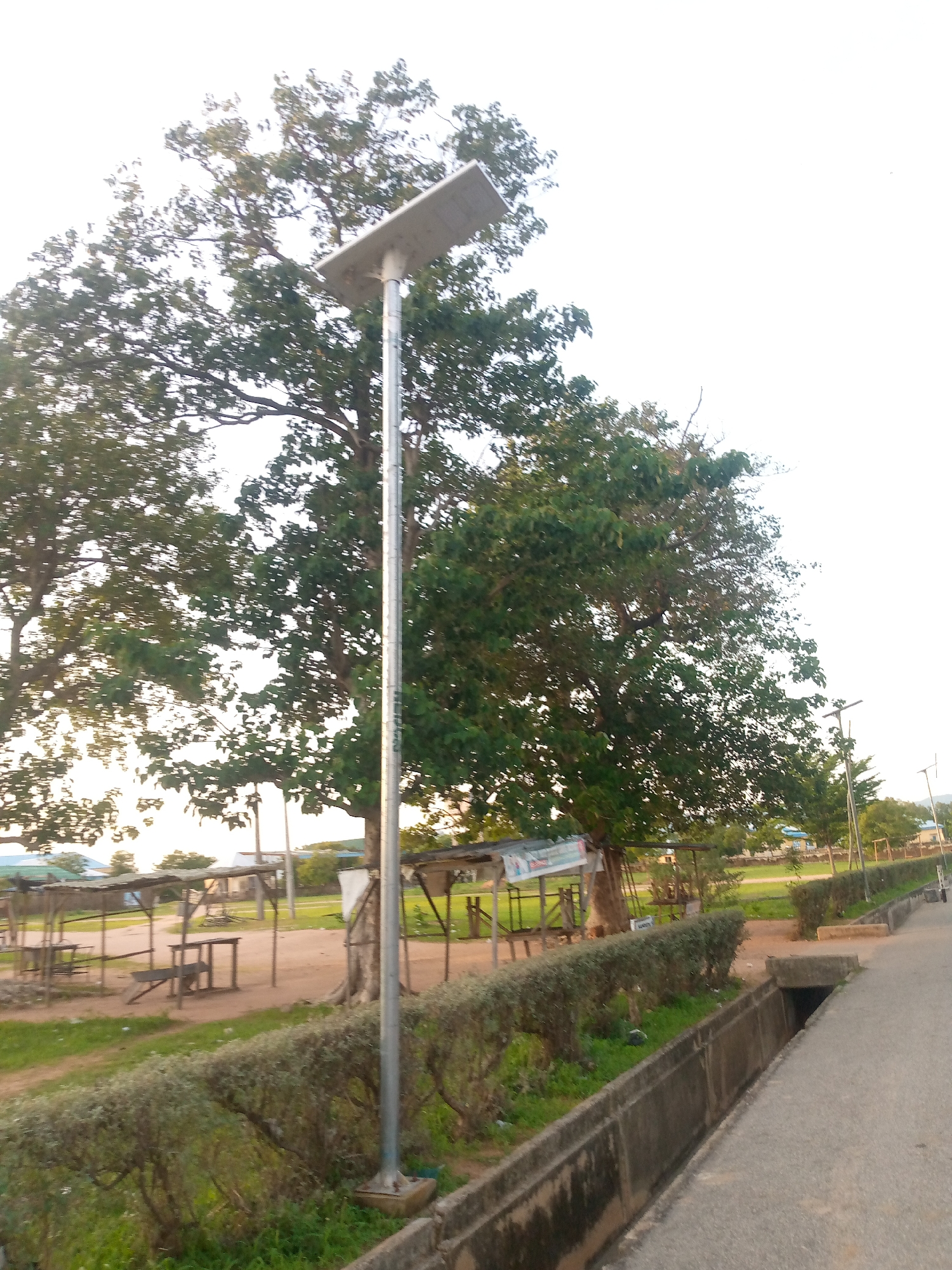 The image depicts an outdoor scene in Jalingo, Nigeria, likely on the outskirts of the city. Dominating the foreground is a tall, slender metal pole topped with a solar panel. Adjacent to the pole, a concrete drainage channel runs alongside a paved road. The channel appears aged and has some vegetation growing within it. Beyond the road and drainage, a neatly trimmed hedge separates the road from an open, sandy area. In this sandy area, there are several open-sided shelters with wooden posts and corrugated metal roofs, some of which appear weathered. Beneath these shelters, wooden benches and tables are visible, suggesting a resting or gathering area. Large, mature trees with dense green foliage are scattered throughout the scene, providing shade. In the background, more trees and some low-lying buildings with blue roofs can be seen, indicating a more developed area further in the distance. The sky is overcast and bright, suggesting daytime. The overall impression is one of a semi-rural or developing urban environment, possibly a rest stop or a community area. There are no people or animals visible in the image.