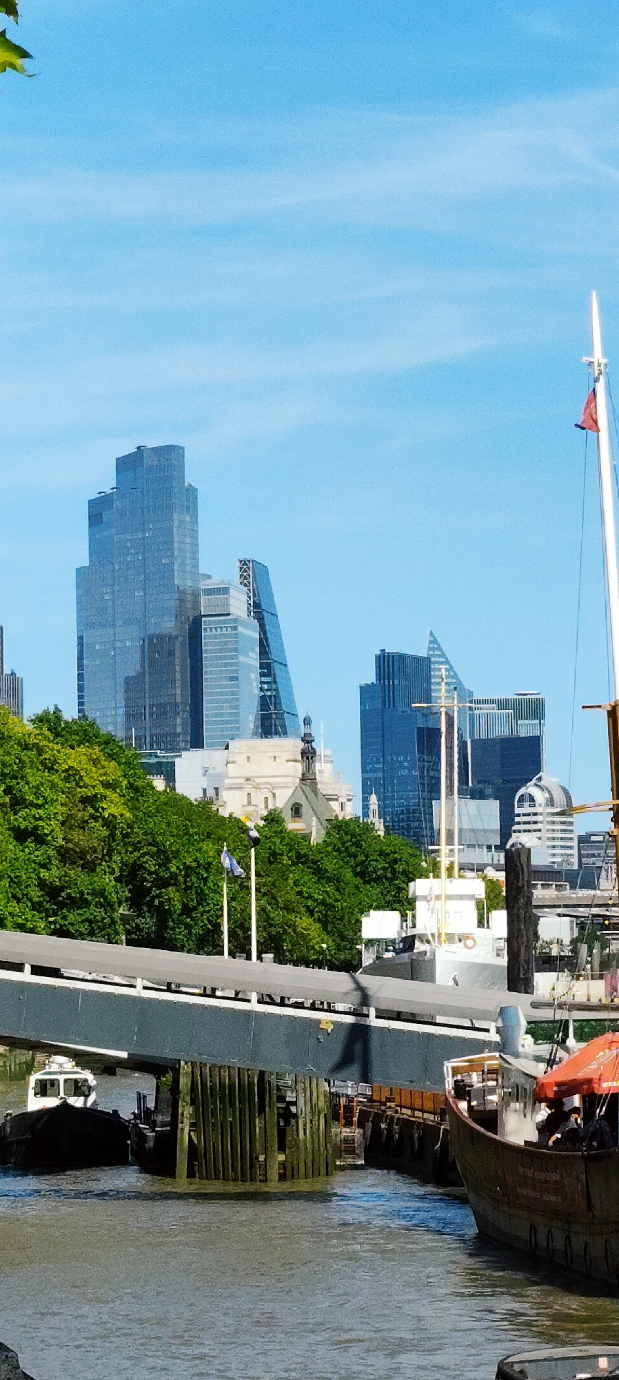 This image captures a waterfront view of the London skyline, featuring modern glass skyscrapers and a vibrant green treeline along the Thames River. In the foreground, a section of a bridge or walkway extends across the scene, with wooden pilings visible in the water beneath it. Two boats are docked or moving in the river; one is a dark vessel with a white cabin, and the other is a larger, older wooden boat with an orange awning. The sky is clear and blue with wispy clouds. In the mid-ground, behind the trees, a white building with architectural details and a steeple is visible, likely a church or historical landmark. The towering glass buildings dominate the background, showcasing contemporary architecture. A flagpole with a red flag is also visible on the right side of the image. The scene appears to be set on a bright, sunny day.