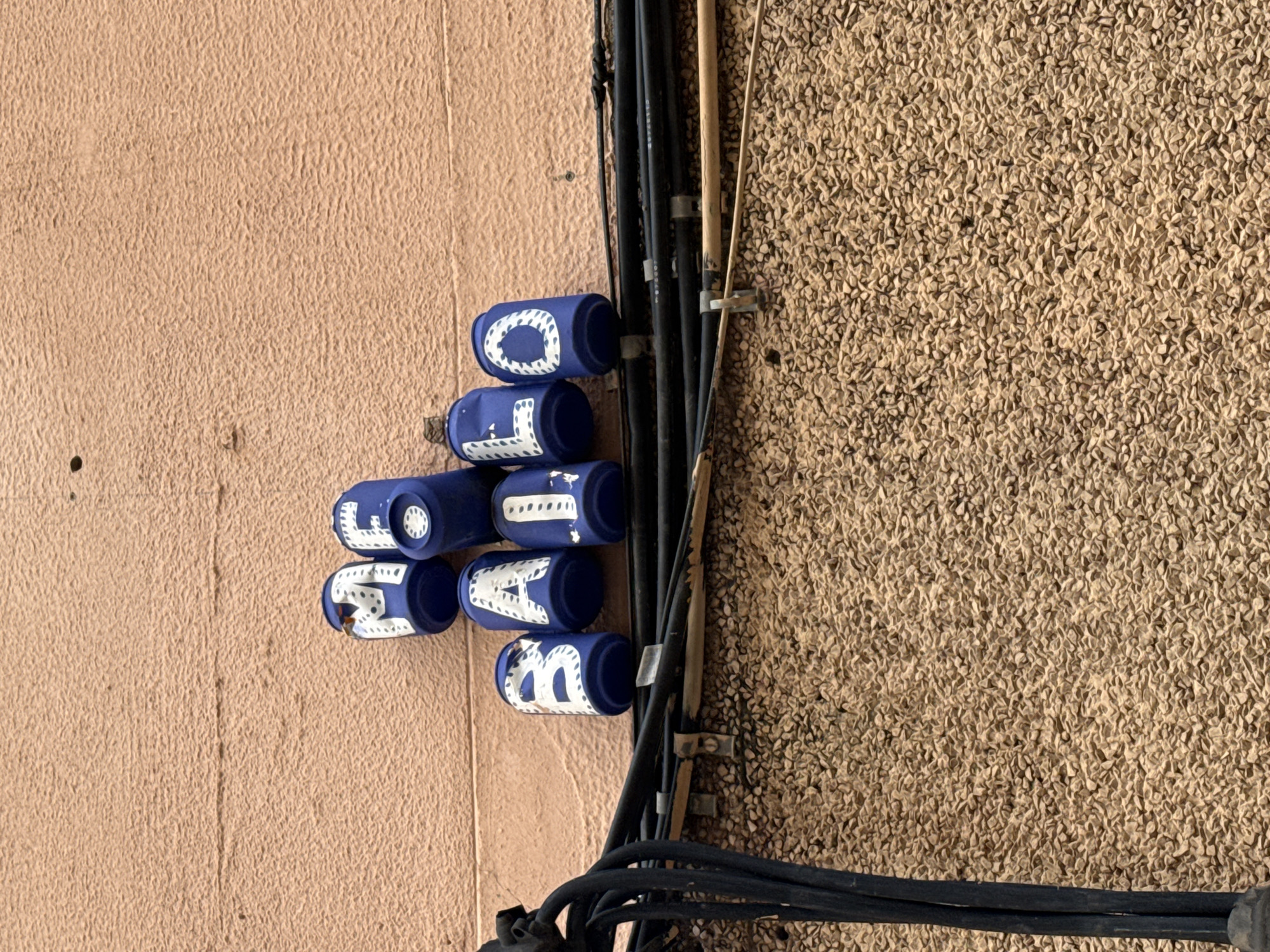 A utility pole is visible on a building facade. The utility pole has several blue caps covering wires and the caps spell out "OLOT MA", possibly indicating the location. The wires are also secured to the building with metal clips. The building has two distinct facade surfaces, one smooth with a stucco finish and a more textured surface made of aggregate material. This is in Barcelona, Spain.