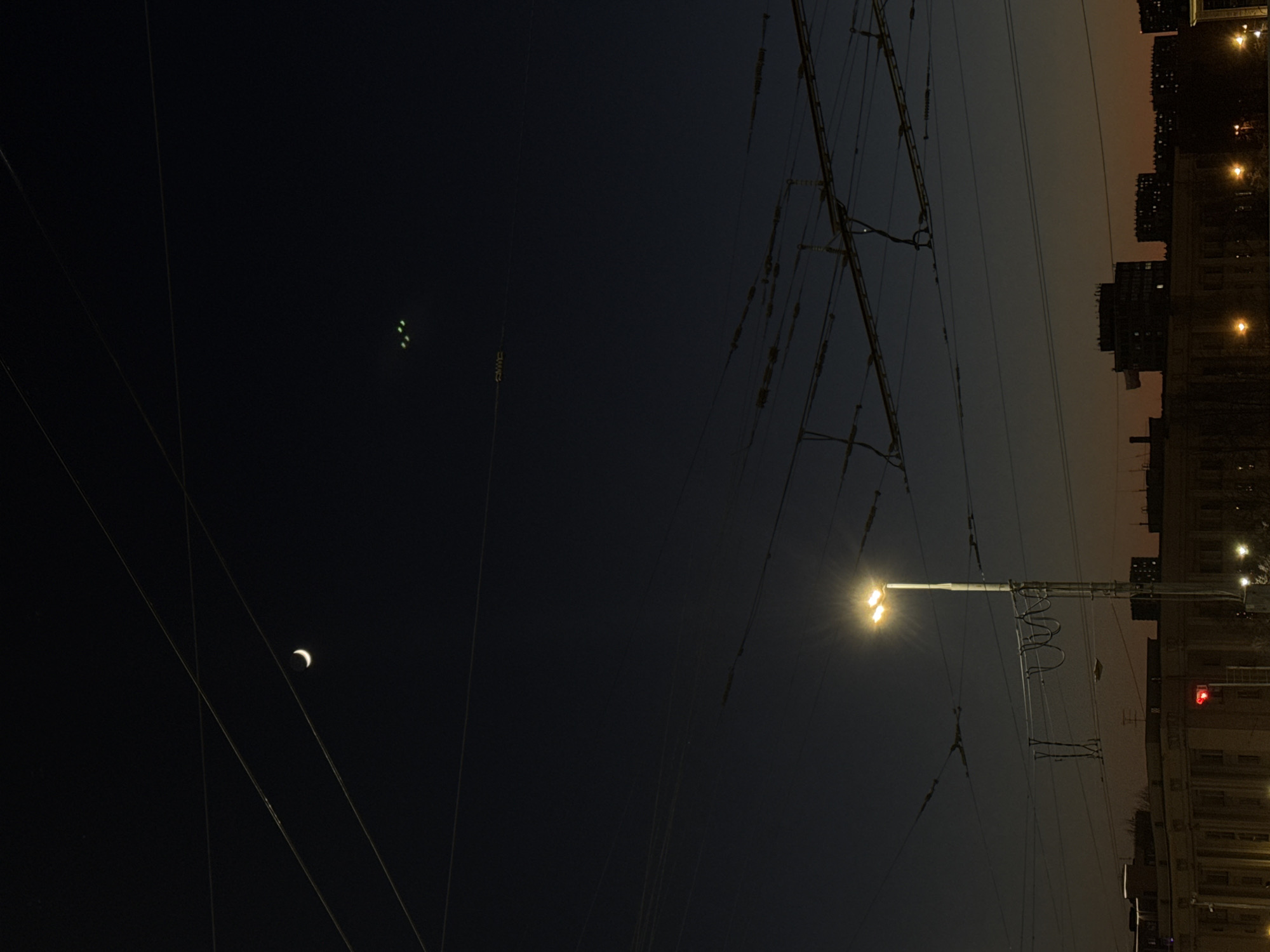 This nighttime photograph, taken in Saint Petersburg, Russia, presents an urban landscape under a dark sky. The image features a complex network of overhead wires, likely for public transport like trolleybuses or trams, crisscrossing the frame. In the upper left quadrant, a distinct crescent moon is visible, and further to its right, three small, faint green lights appear in the dark sky, possibly originating from an aircraft or distant city lights.
On the right side, a prominent street light pole with two bright yellow lamps illuminates the scene, casting a strong lens flare. Below and extending to the right, the silhouettes of multi-story urban buildings form the horizon, with subtle internal lights visible in some windows. A red traffic light is partially visible on the far right, situated behind the main streetlight pole.
The sky transitions from a deep, dark blue at the top to a slightly lighter, warmer orange-brown hue near the buildings, suggesting either atmospheric conditions at twilight or the effects of urban light pollution. The overall impression is one of a quiet, illuminated city evening. The weather appears clear, allowing for visibility of both celestial and artificial lights. No specific human activity or visible text is discernible within the frame.