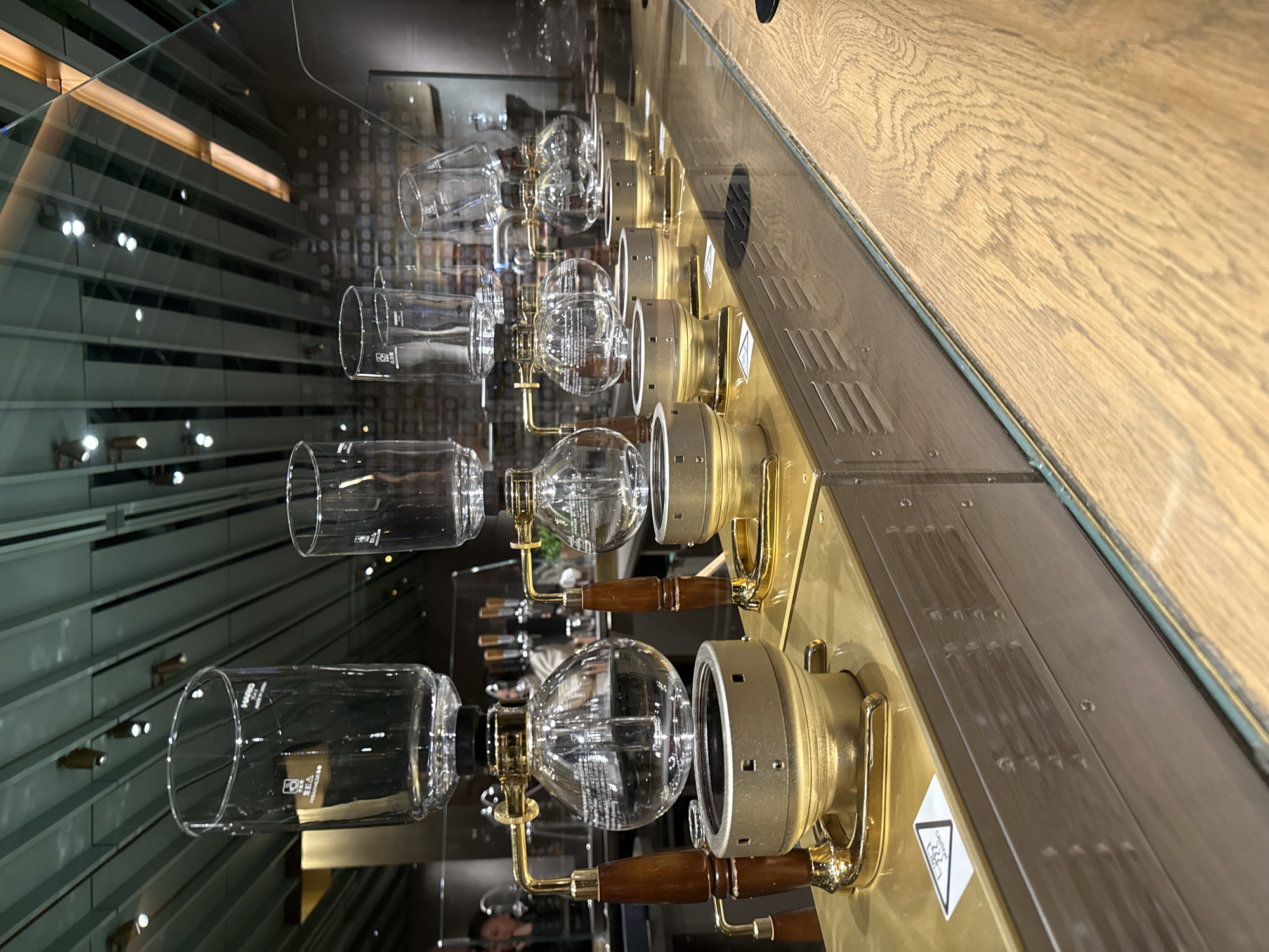 The image shows a row of four siphon coffee brewers, each consisting of a round glass boiling flask below and a cylindrical glass brewing chamber above, connected by a narrow tube. The brewing apparatus is mounted on a metal base with wooden handles. The background, visible through a glass partition and reflections, suggests a modern interior, potentially a cafe or restaurant. The location can be inferred to be the Starbucks Reserve Roastery in Chicago, United States of America, based on the style of the coffee brewing equipment and the architectural details. There are no people visible in the immediate foreground, though faint reflections indicate their presence nearby.