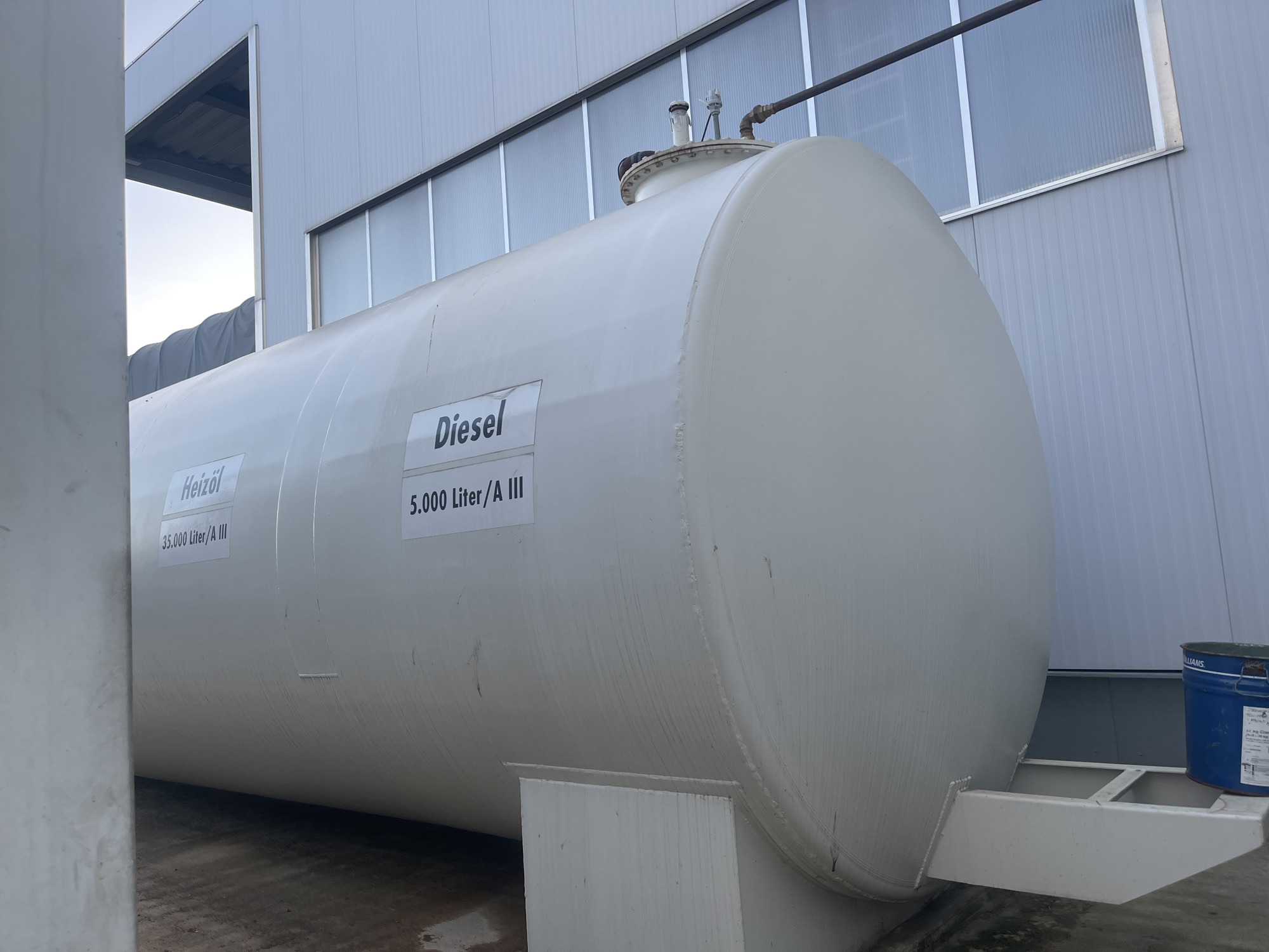 The image depicts a large, horizontal, cylindrical tank located outdoors. The tank is painted white and has two labels affixed to its side. The first label reads "Heizöl 35.000 Liter/A III" and the second reads "Diesel 5.000 Liter/A III". The tank is supported by a rectangular base and has various fittings on top. Behind the tank, there is a light gray building with a series of windows along the top portion of the wall. The environment appears to be a paved or concrete area. A blue container with a label is partially visible on the right side of the image. The specific location is Unknown, Unknown.