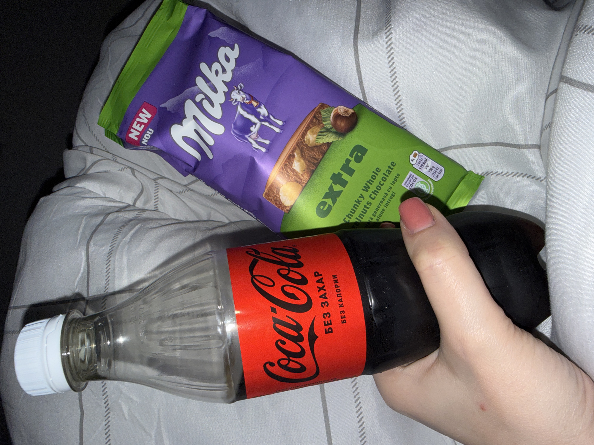 The image depicts an indoor scene within the city of Varna, Bulgaria. A pale-skinned hand, exhibiting short, light-pink manicured fingernails, firmly grips a clear plastic Coca-Cola bottle containing a dark brown liquid. The bottle's label is predominantly red, displaying the "Coca-Cola" logo in white script, with additional Cyrillic text below: "БЕЗ ЗАХАР" (sugar-free) and "БЕЗ КАЛОРИИ" (calorie-free). The bottle is sealed with a white, ribbed plastic screw cap. Adjacent to the Coca-Cola bottle, a purple and green flexible wrapper for a "Milka extra Chunky Whole Hazelnuts Chocolate" bar is visible. The wrapper features the "Milka" logo, a depiction of a white and purple cow, and an image of the chocolate bar with hazelnuts. Text such as "NEW NOU" and "SUSTAINABLY SOURCED COCOA," alongside Romanian text "cu lapte alune întregi," is also visible on the packaging. Both items are positioned on or against a background of light-colored bedding, characterized by subtle, intermittent grey horizontal stripe patterns, consistent with a duvet or sheet in a bedroom setting.
