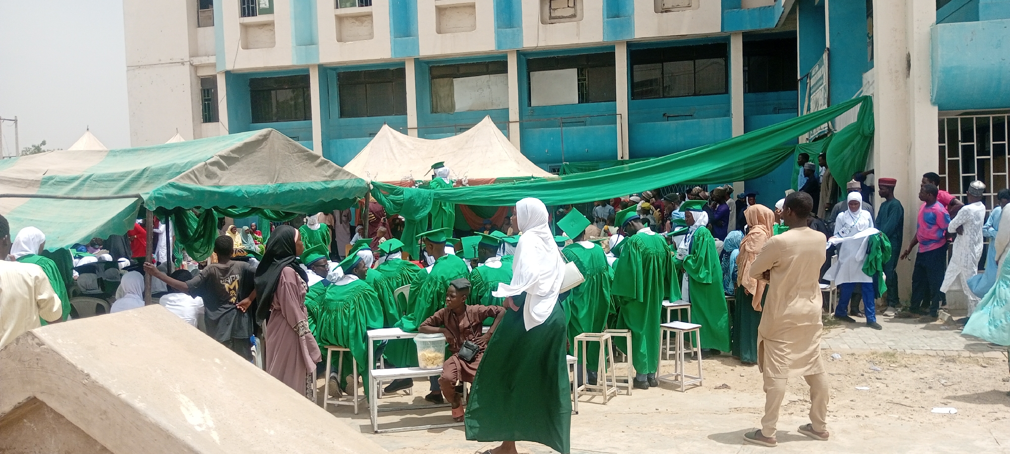 Captured in the city of Gwagwarwa, Nigeria, the image shows an outdoor graduation ceremony. The event appears to be taking place on the grounds of a multi-story building with a blue and white facade. Several large tents are set up to provide shade for attendees, and green fabric drapes from the tents and building. A diverse group of people are present, many of whom are wearing green graduation gowns and caps, seated on rows of white plastic chairs. Some attendees are dressed in traditional Nigerian clothing, and others wear Western attire. A person in a white headscarf and green dress stands prominently in the foreground. Other individuals are scattered around the perimeter of the seating area, some standing, some sitting, and others interacting with one another.