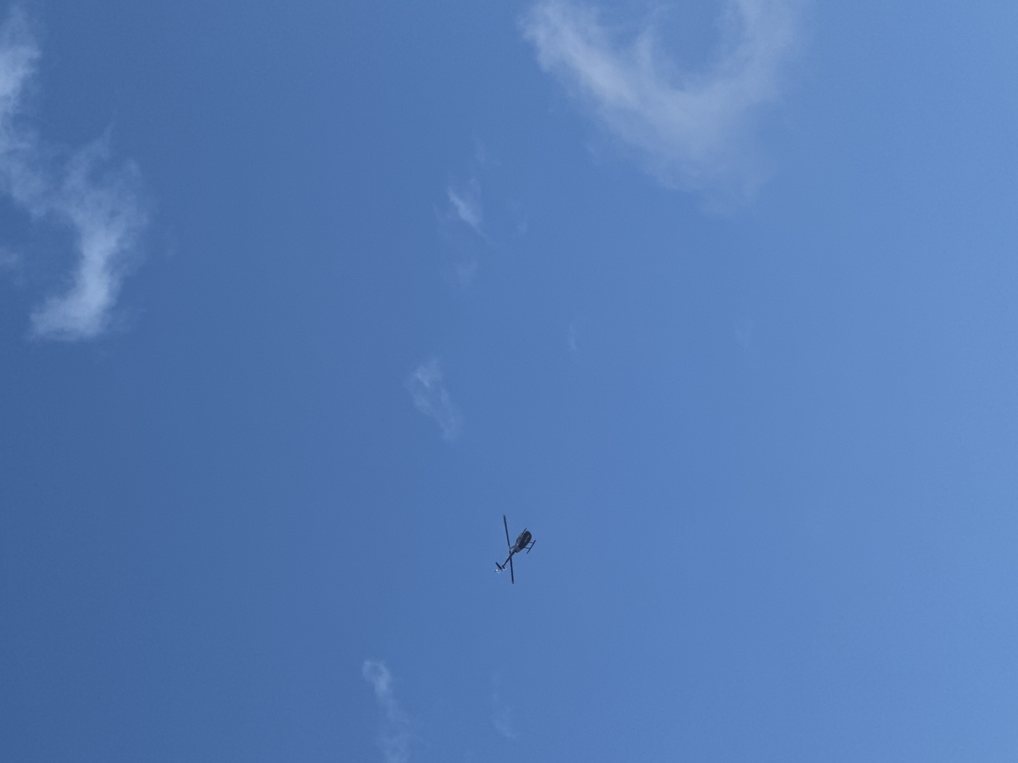 A solitary dark-colored helicopter is captured in mid-flight against a clear blue sky with scattered wispy clouds. The helicopter appears to be a smaller model, with its rotors visible in motion. The image was taken in Kuta Selatan, Indonesia.