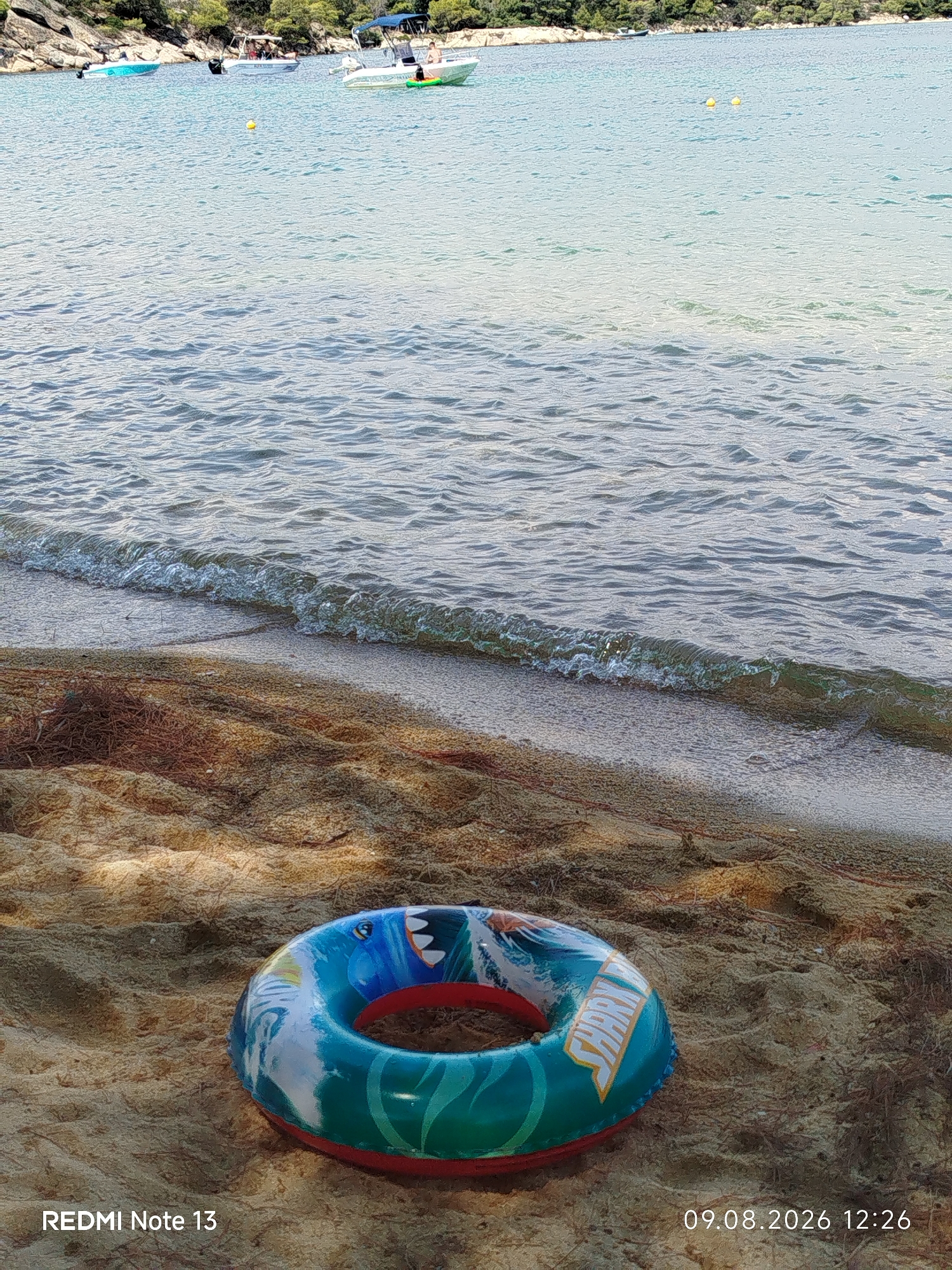 An inflatable ring with a shark design rests on the sandy shore of a beach in Sithonia Municipal Unit, Greece, with the gentle waves of the Aegean Sea lapping at the sand in the background. The water is a clear blue, and several boats are visible in the distance, suggesting a sunny day. The beach is sparsely populated, with a few people visible on one of the boats. The image appears to have been taken on August 9, 2026, at 12:26 PM.