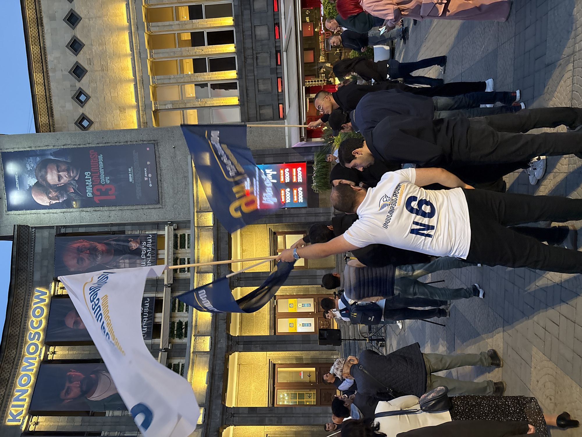 The image shows a group of people gathered outside the "KINOMOSCOW" cinema in Yerevan, Armenia. Some individuals are lying down on the sidewalk. Several people are holding flags, including one that is white and another that is blue. A man in the foreground is wearing a white t-shirt with the number "N6" on the back. Movie posters are visible on the facade of the cinema building. A large sign with flashing numbers and text is also visible behind the group.